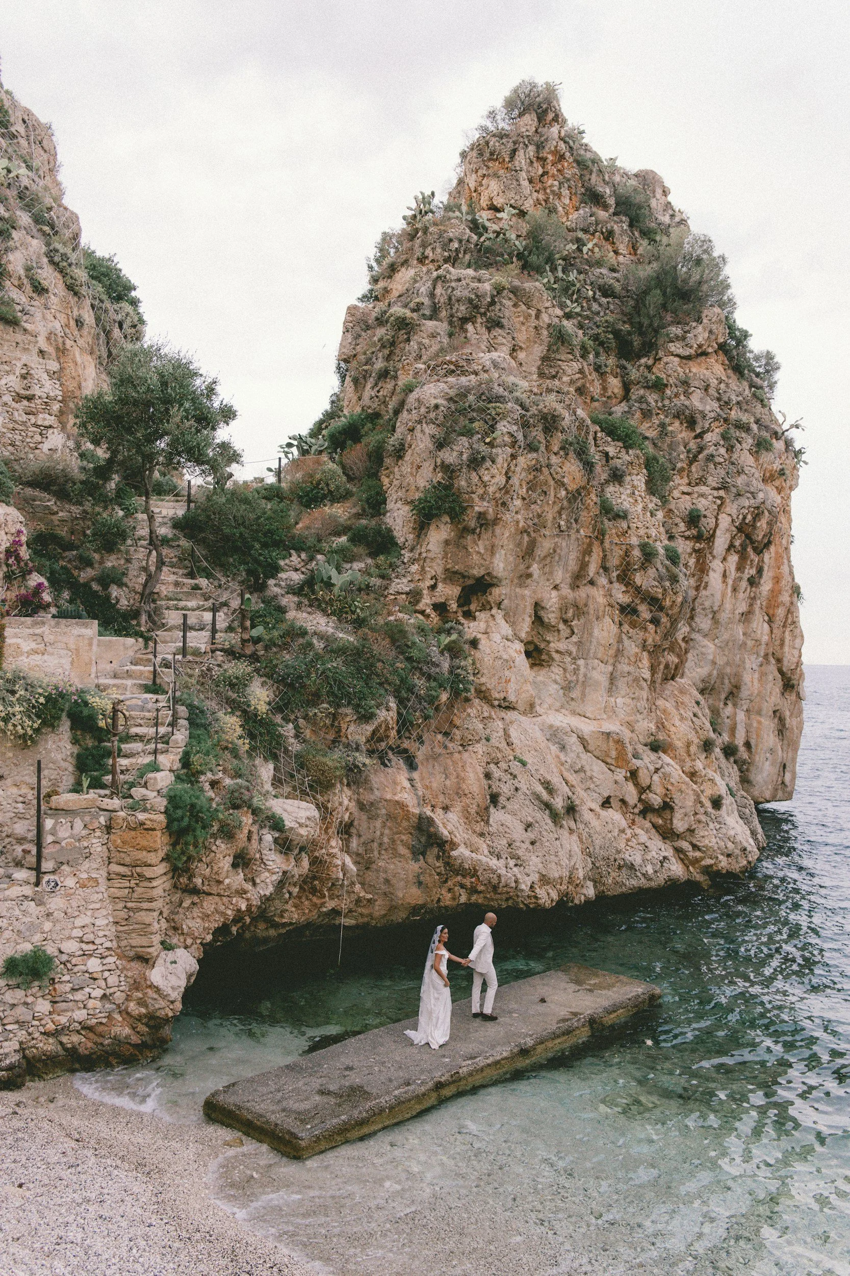 A couple dressed in wedding attire stand on a concrete platform at the beach, holding hands, with a large rocky cliff and stairs with plants and trees behind them.