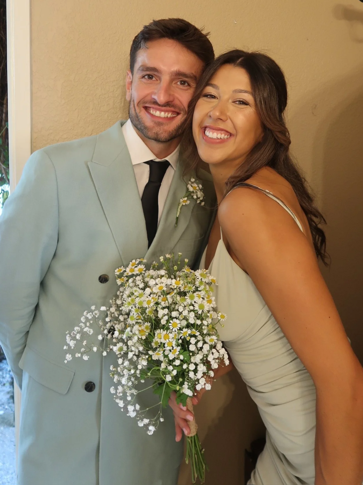 A smiling couple, dressed in wedding attire, standing close together with the bride holding a bouquet of wildflowers.