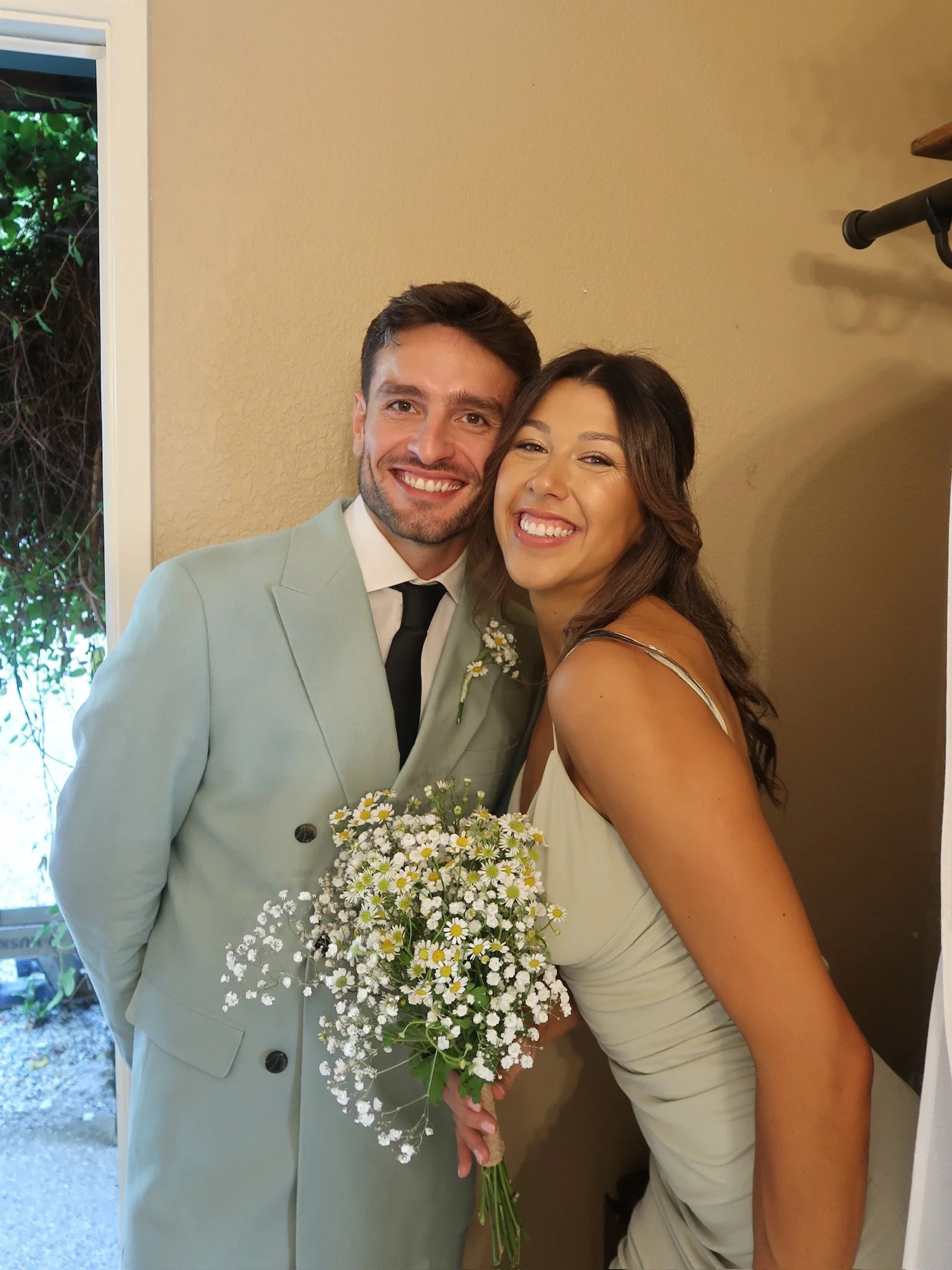 Smiling man and woman on a wedding day, the man wearing a light green suit and black tie, holding a bouquet of white daisies with yellow centers, standing near a doorway.