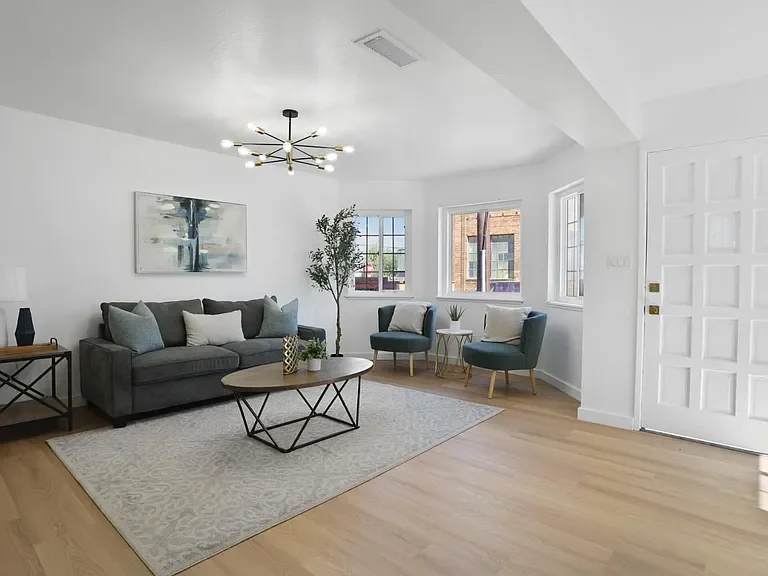 Bright living room with white walls, wooden floor, gray sofa, two teal armchairs with pillows, a wooden coffee table, a patterned area rug, modern chandelier, artwork on the wall, window with city view, houseplants, and a white door.