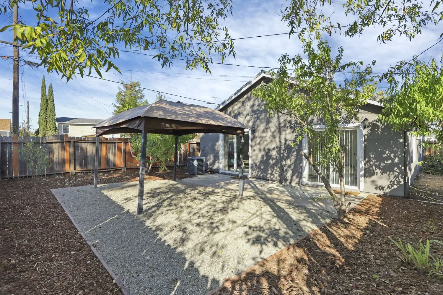 Backyard with a small gravel patio area, a patio cover, a tree, and a house with sliding glass doors.