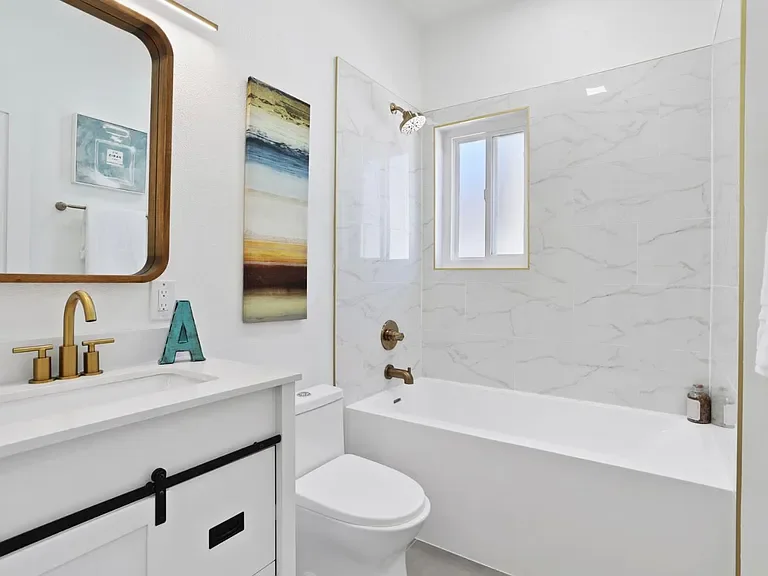 A modern bathroom with a white bathtub, a toilet, a vanity with a gold faucet, a mirror, and art on the wall. There is a window above the bathtub, and the walls are tiled with white marble.