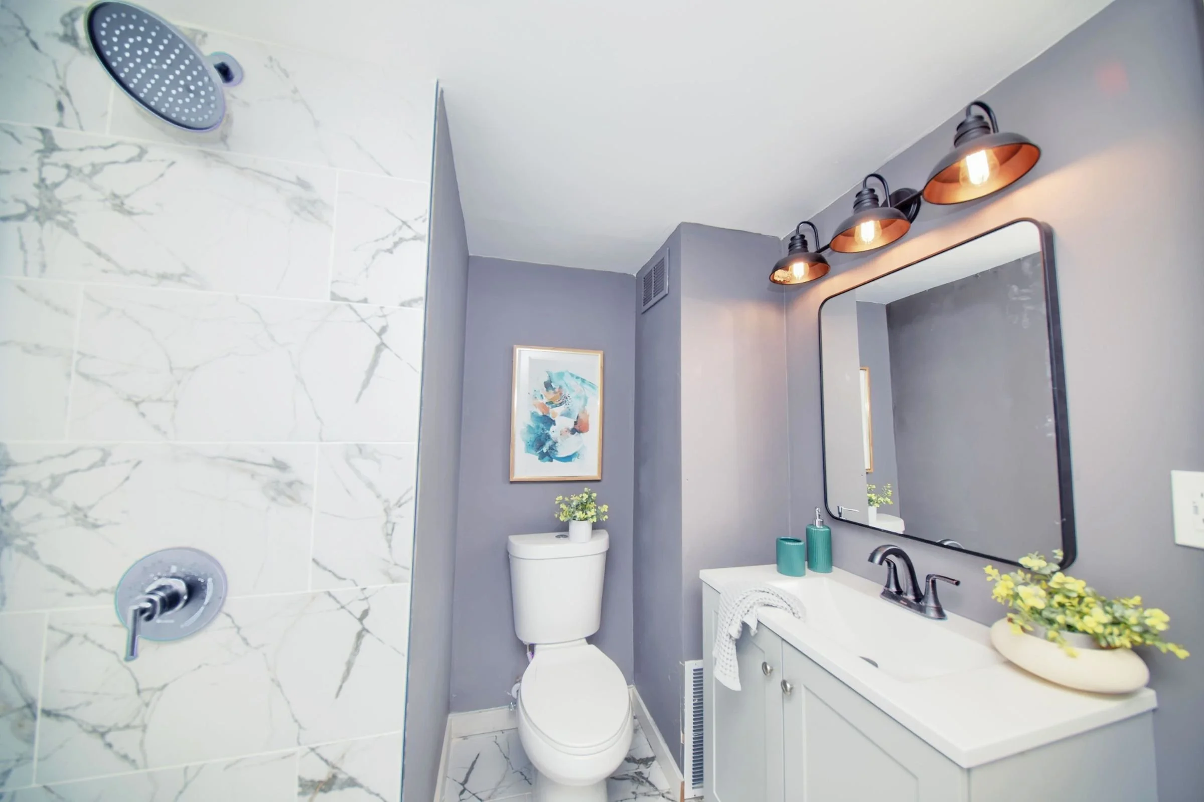 Bathroom with marble shower on the left, toilet with a plant on top, and a vanity with a mirror, plants, and towels. Gray walls and black fixtures, decorated with framed artwork.