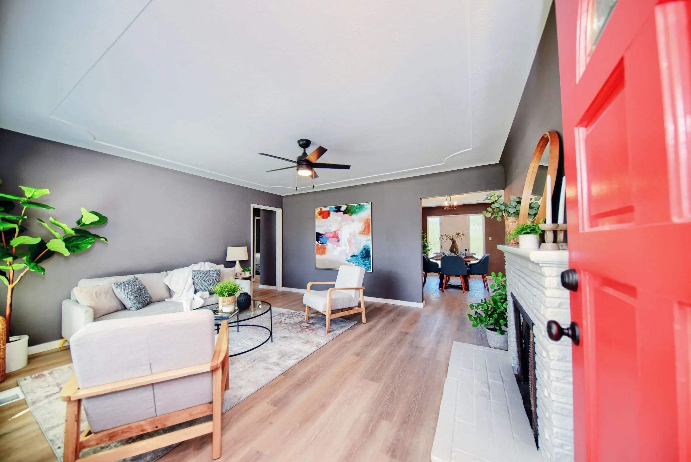 Living room with gray walls, white couch, white armchair, black ceiling fan, colorful abstract art on wall, fireplace with white brick surround, and a red door partially open in the foreground.
