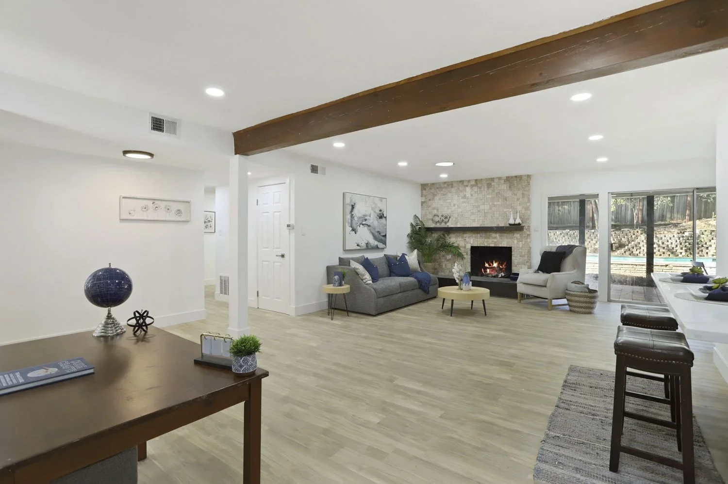 Living room with white walls, a gray sofa with navy pillows, a white armchair near glass sliding doors, a fireplace with a stone mantel, wooden beams on the ceiling, and a view of an outdoor pool area.