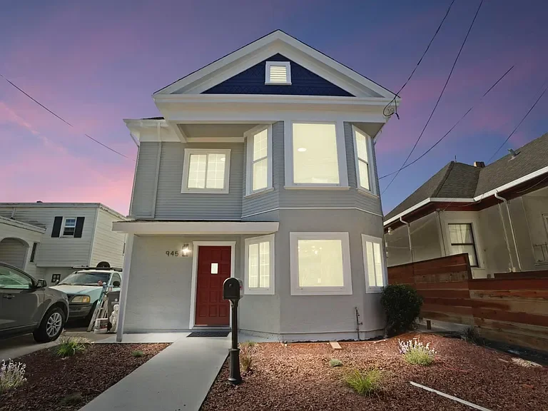 Two-story house with grey exterior, red front door, multiple large windows, and a small front yard with mulch and plants, during sunset.