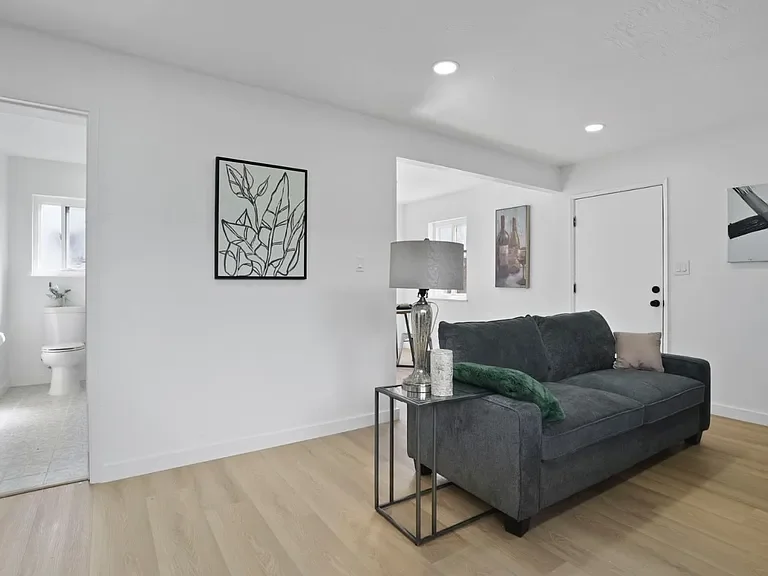 Living room with a dark gray sofa, a side table with a lamp and decorative items, abstract art on the wall, and an open view to a bathroom.