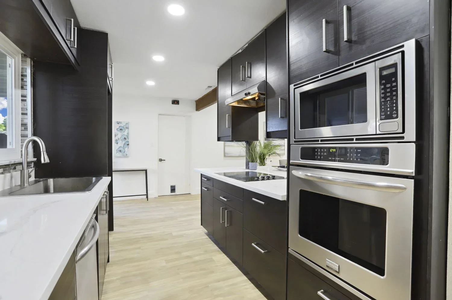 Modern kitchen with black cabinets, stainless steel appliances, white countertops, a window above the sink, and light wood flooring.