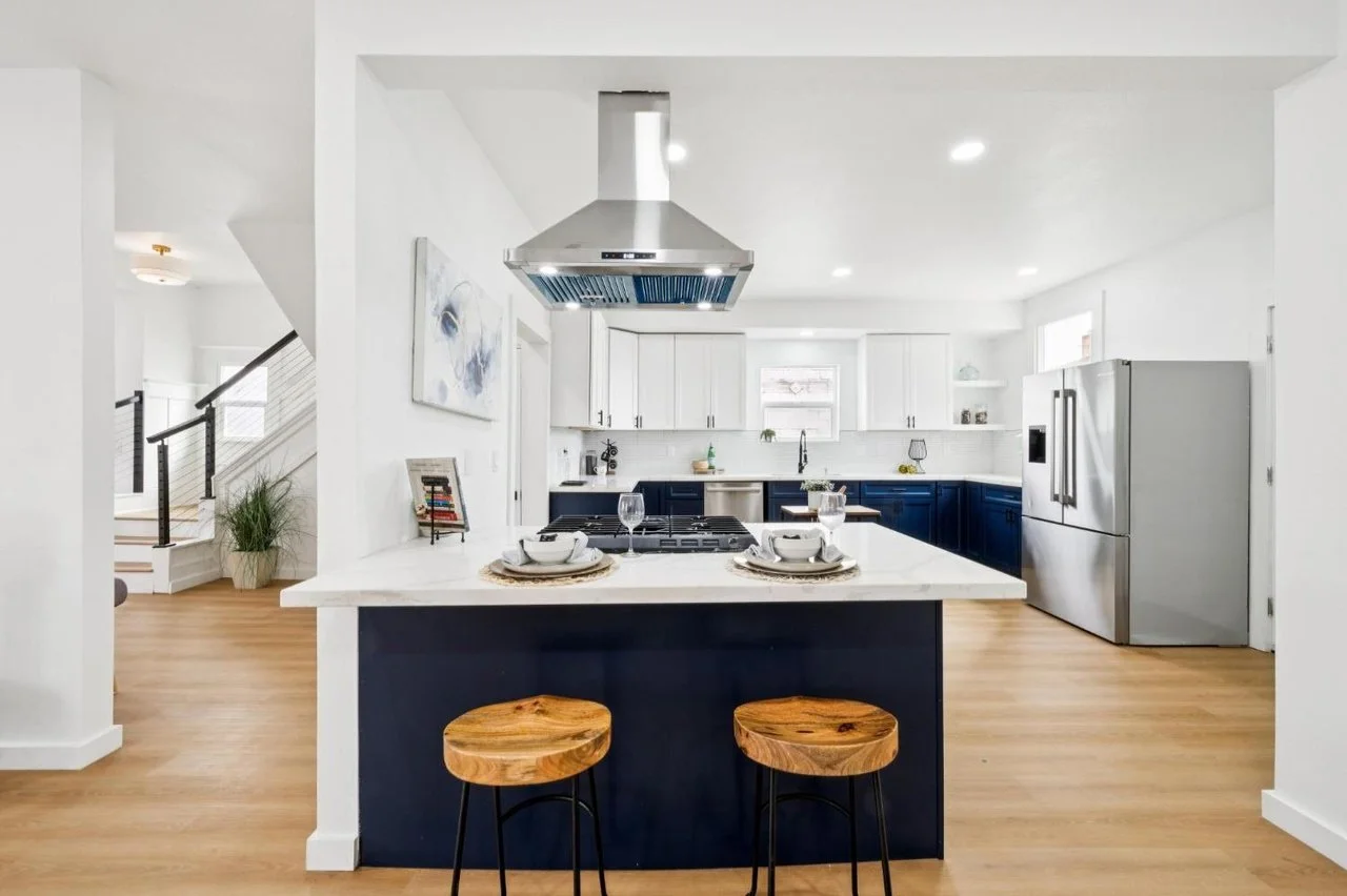 Modern kitchen with white and navy cabinets, stainless steel refrigerator, marble island with two wooden stools, and minimalist decor.