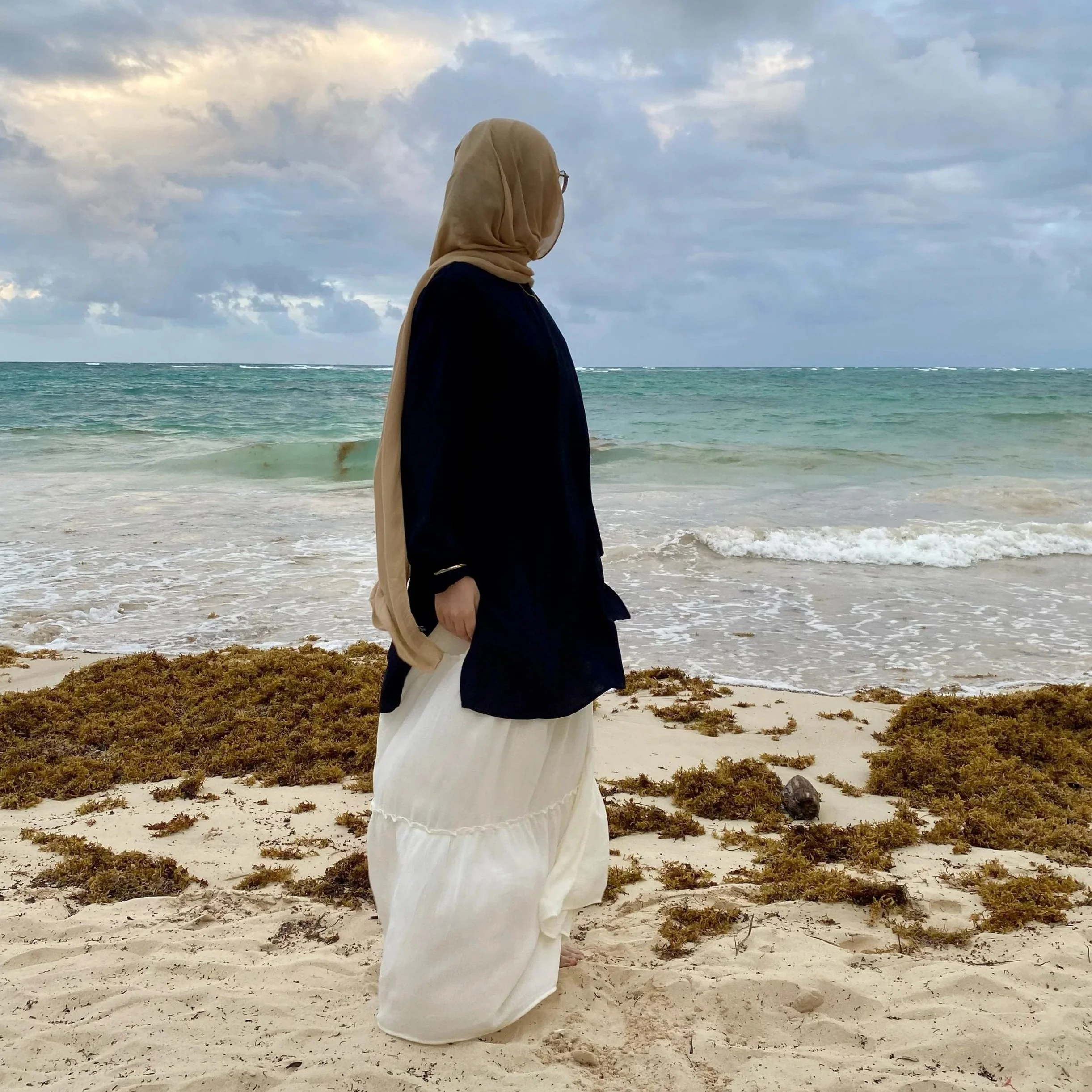 A woman wearing a beige hijab, black blouse, and white skirt, standing on a sandy beach with seaweed, looking toward the ocean under a cloudy sky.