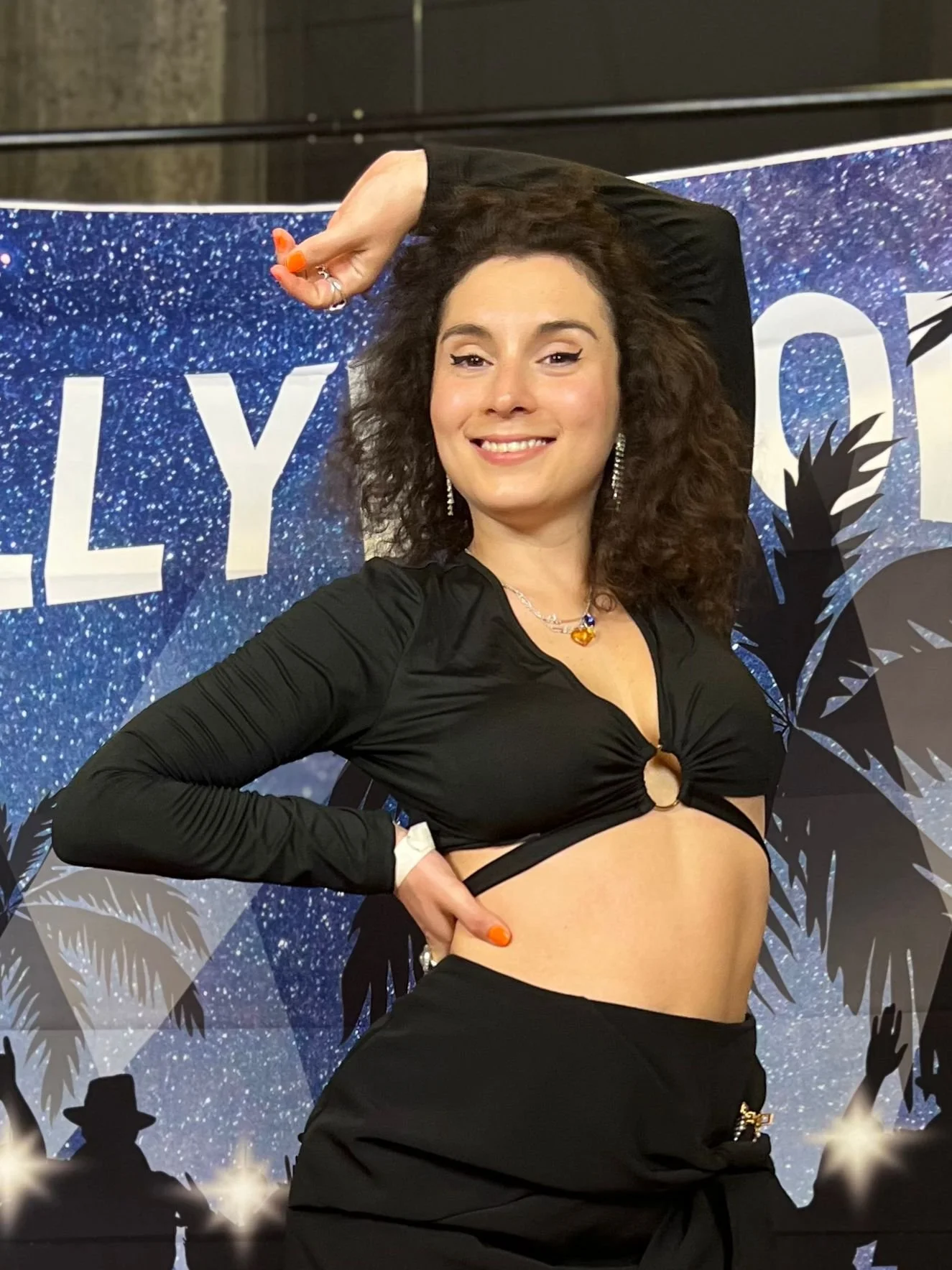 A woman with curly hair smiling and posing with one arm raised and the other on her hip in front of a festive backdrop with the word 'LILY' visible.