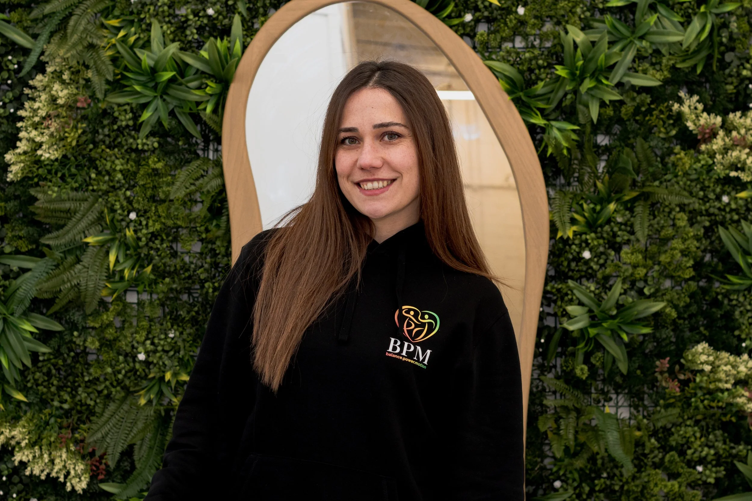 A young woman with long brown hair, smiling, wearing a black top with cutout shoulders and small earrings.