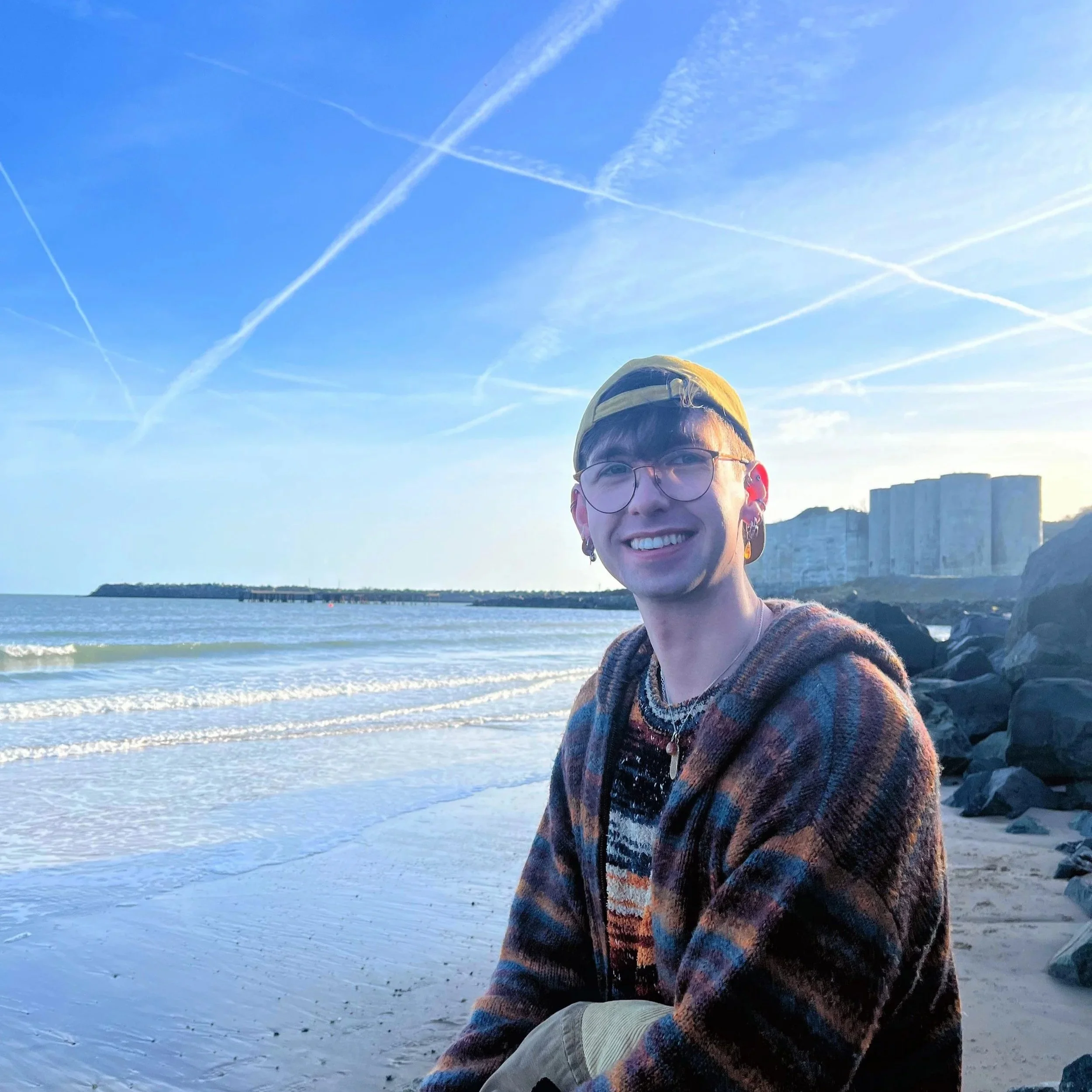 A young person with glasses, earrings, and layered necklaces wearing a yellow cap and a multicolored hoodie, sitting on a sandy beach with rocks, smiling. The background includes the ocean, a pier, and buildings with a blue sky and crisscrossing contrails.