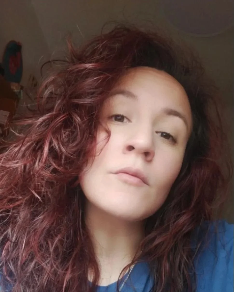 Close-up of a woman with long, curly, reddish-brown hair, looking at the camera with a neutral expression, wearing a blue top.