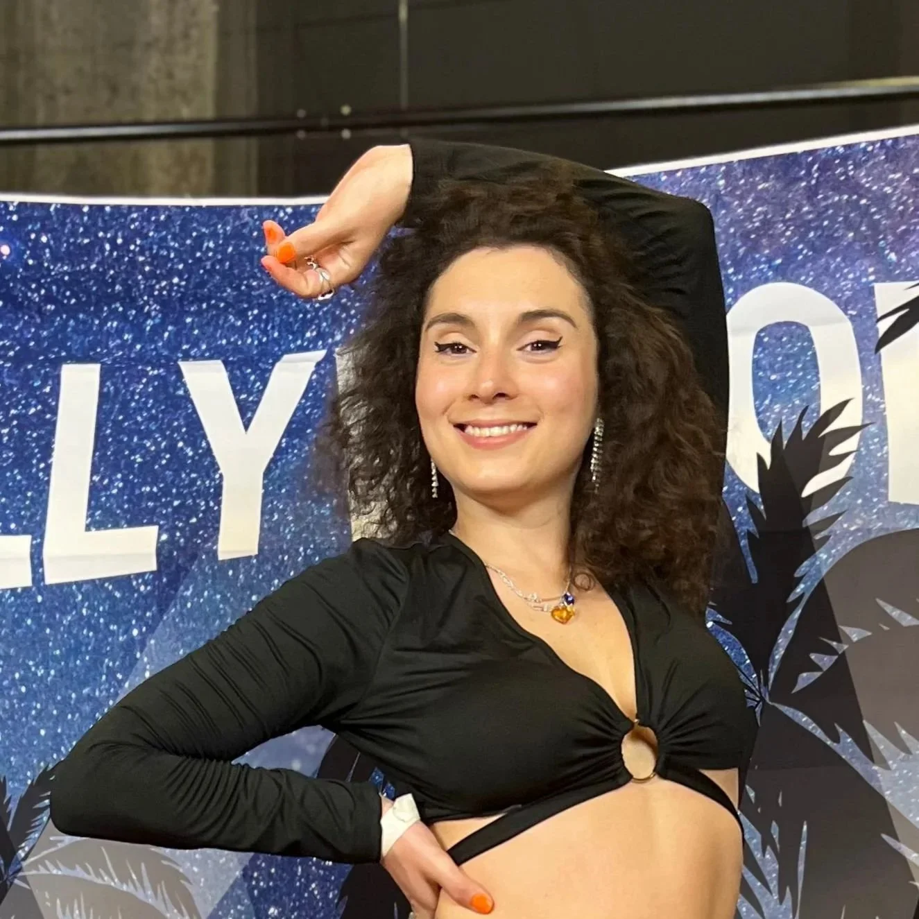 A young woman with curly brown hair smiling, wearing a black long-sleeve crop top, earrings, a necklace, and posing with one arm raised above her head in front of a blue glittery backdrop with the partial word 'LILY' visible.