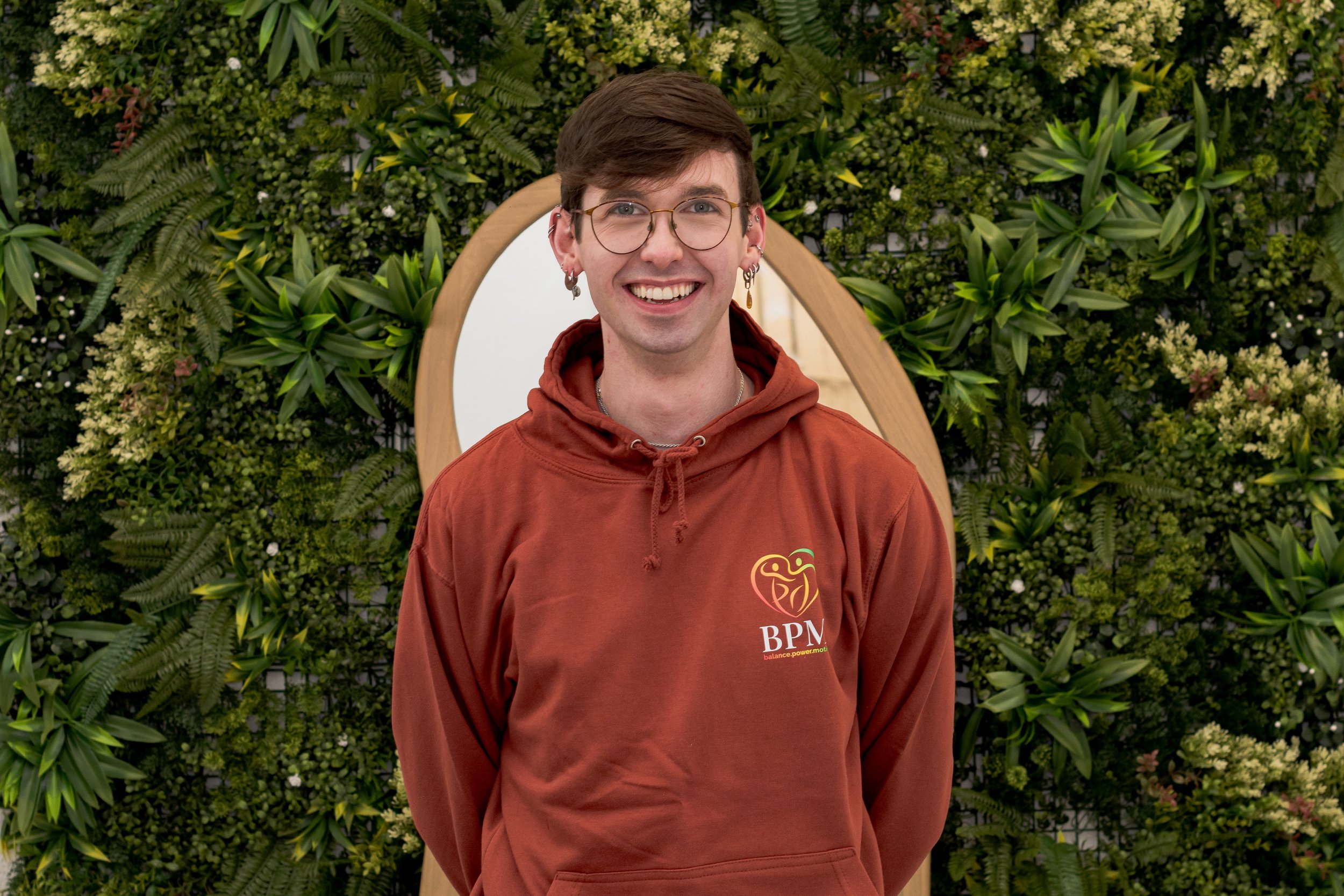 Person sitting on a sandy beach near rocks, smiling, wearing glasses, a yellow cap, and a colorful sweater, with the ocean, shoreline, and buildings in the background on a clear, sunny day.