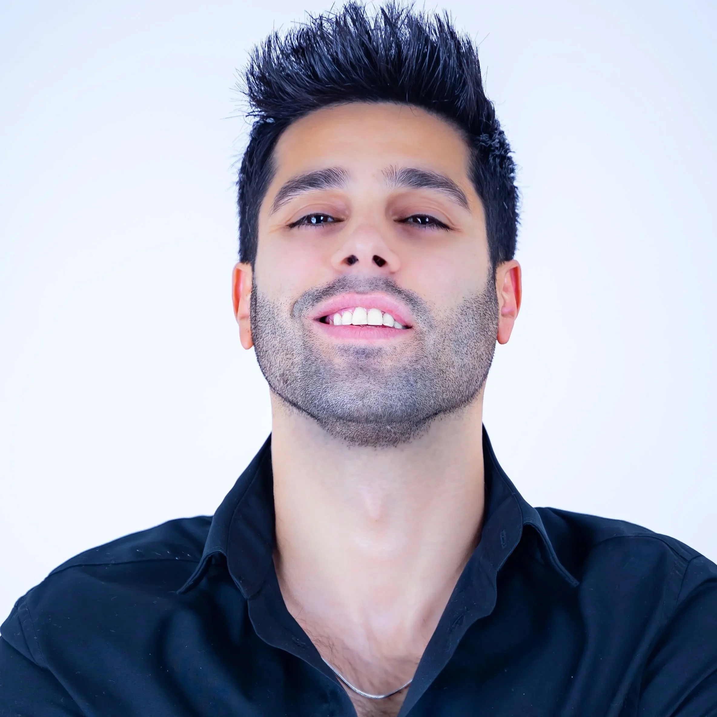 Close-up of a young man with dark, spiked hair, a well-groomed beard, and wearing a black shirt, smiling slightly with a confident expression against a plain light background.