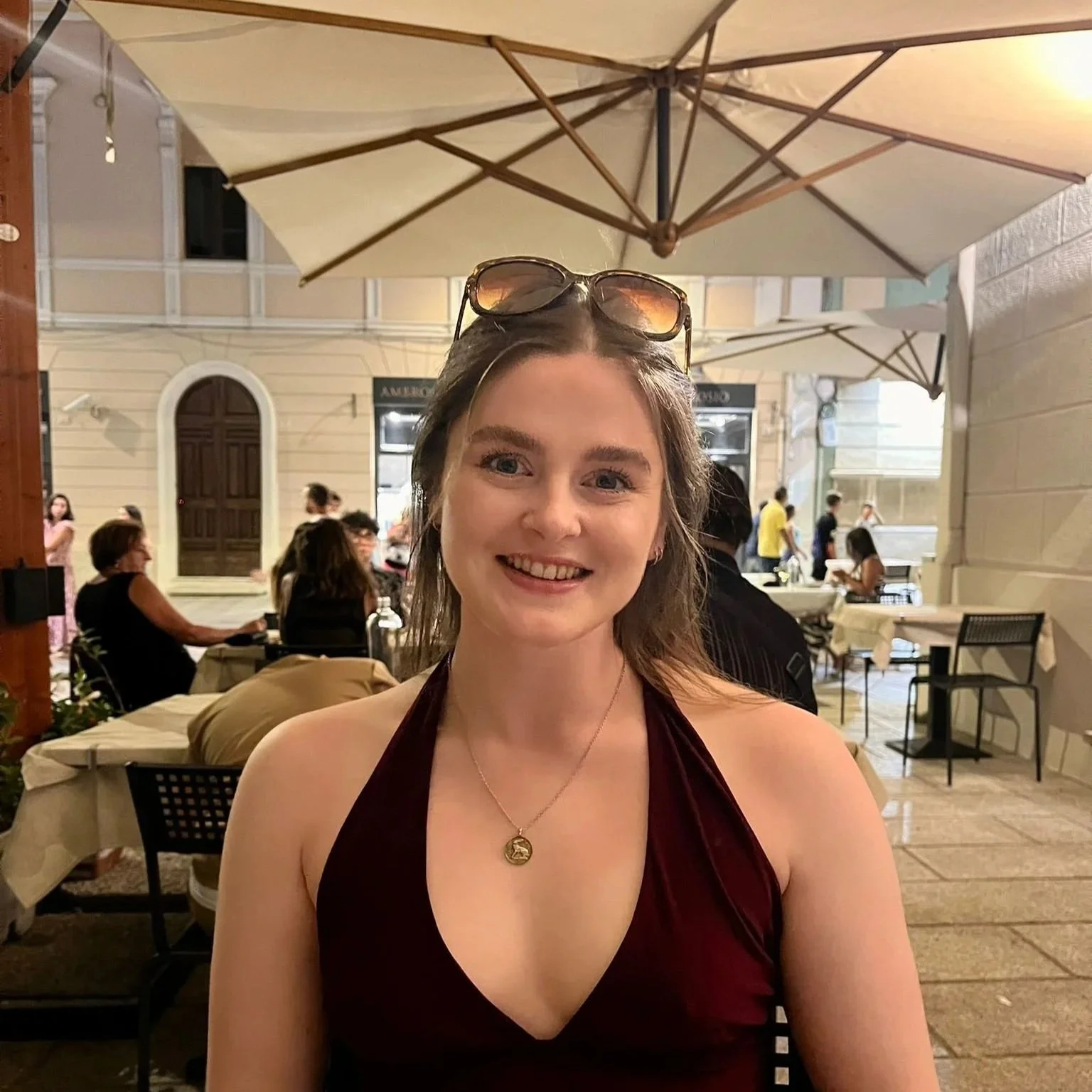 A young woman with glasses on her head, smiling at the camera, sitting at an outdoor restaurant under a large beige umbrella.