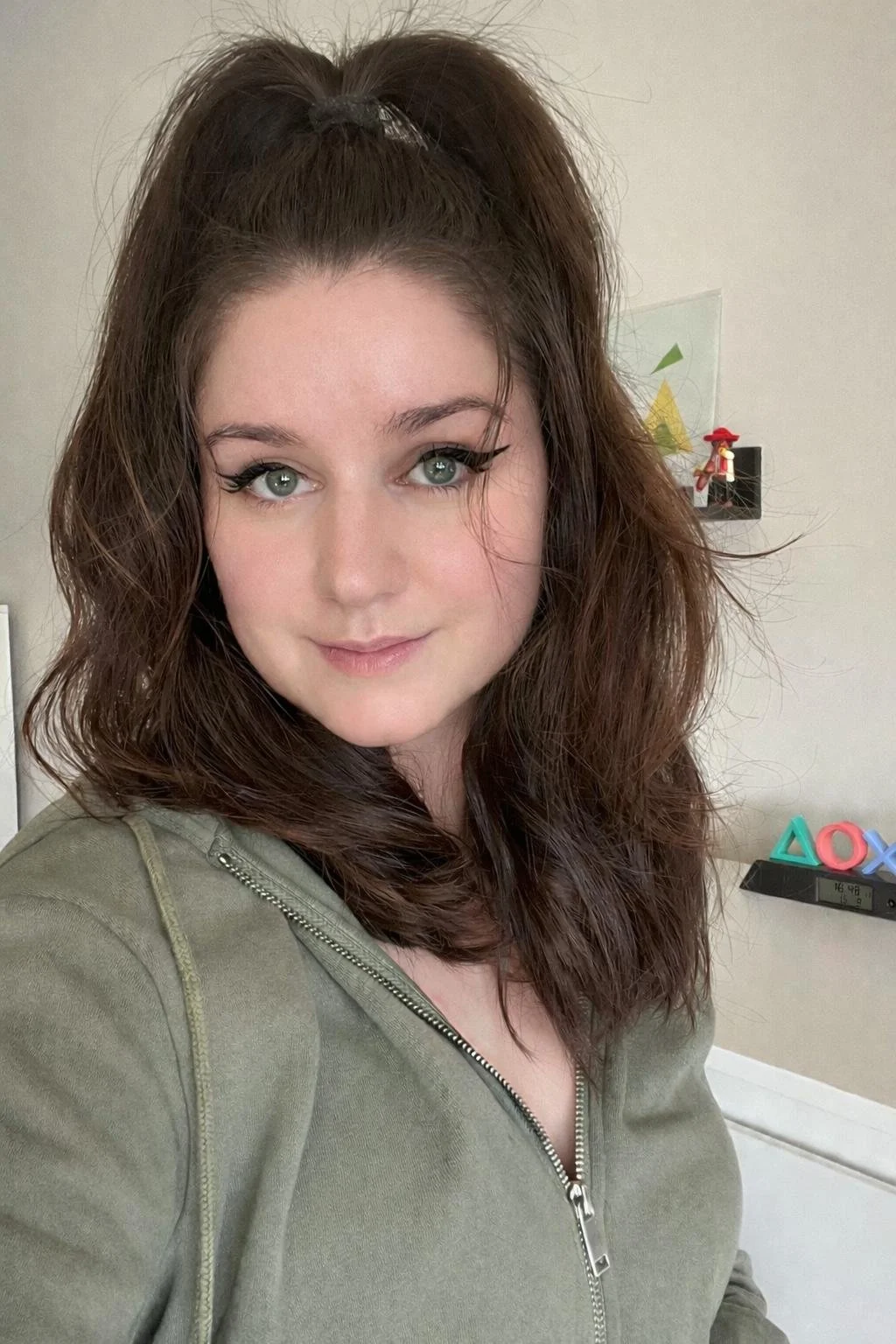 A woman with wavy brown hair styled in a high ponytail, wearing makeup with prominent eyeliner and mascara, taking a selfie indoors.