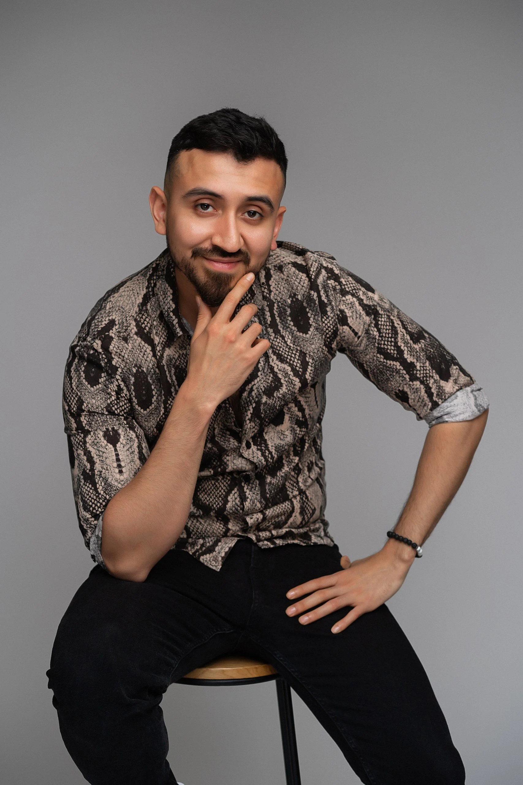 A man sitting on a stool, wearing a snakeskin pattern shirt, with one hand on his chin and the other on his hip, against a plain gray background.