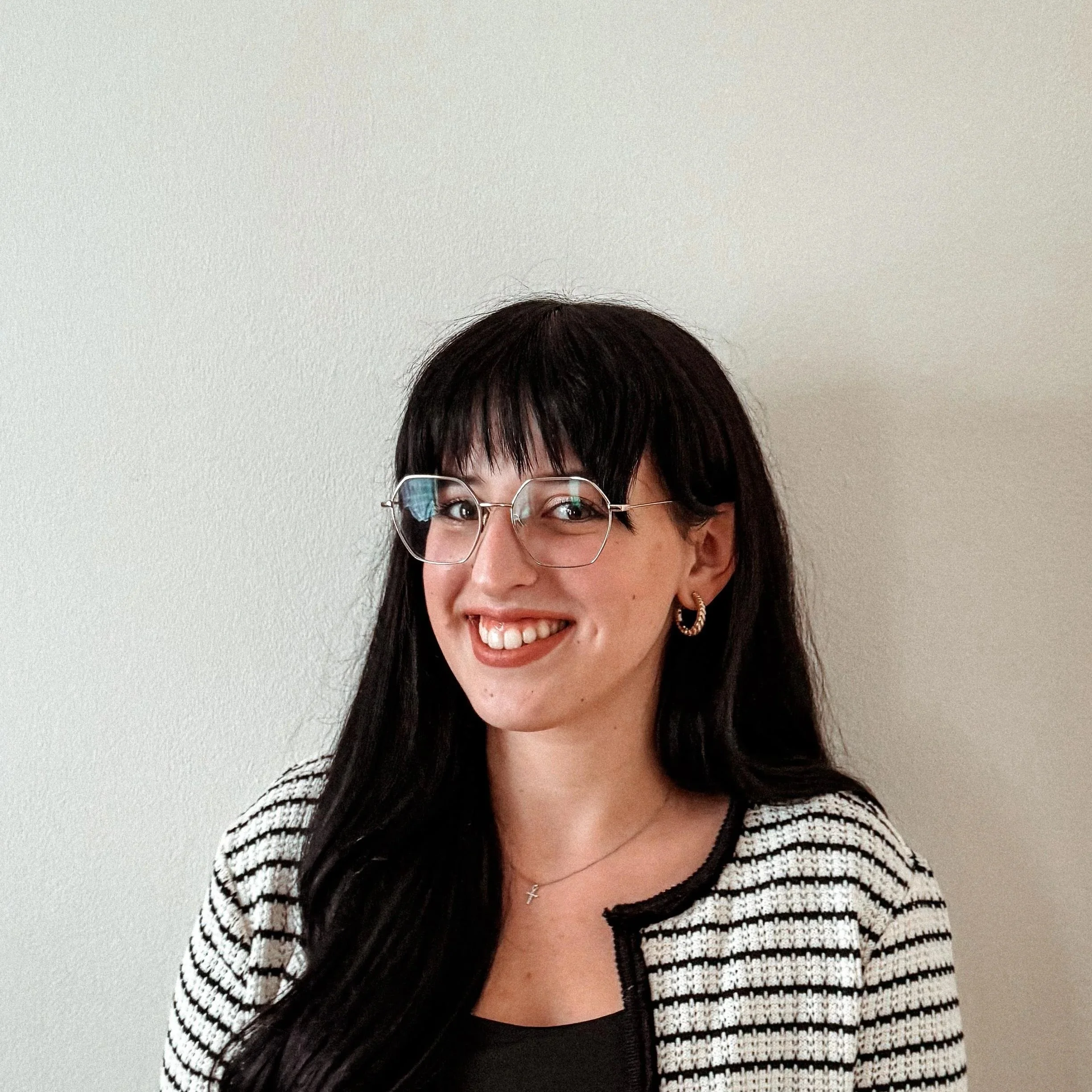 A woman with long dark hair, wearing glasses, earrings, a black top, and a striped blazer, smiling in front of a plain white wall.