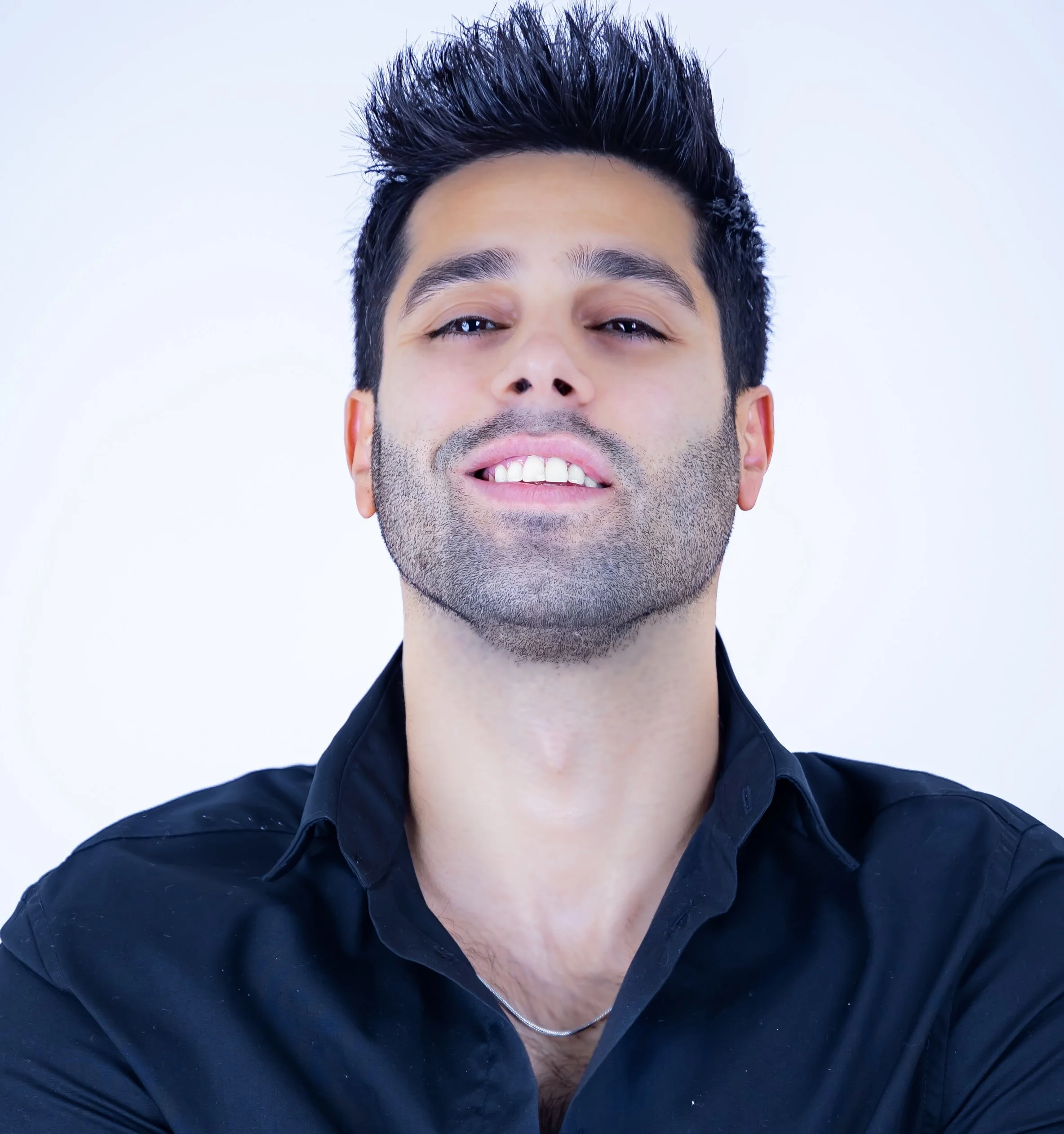 Young man with styled black hair, slight beard, wearing a black shirt, smiling confidently against a light background.