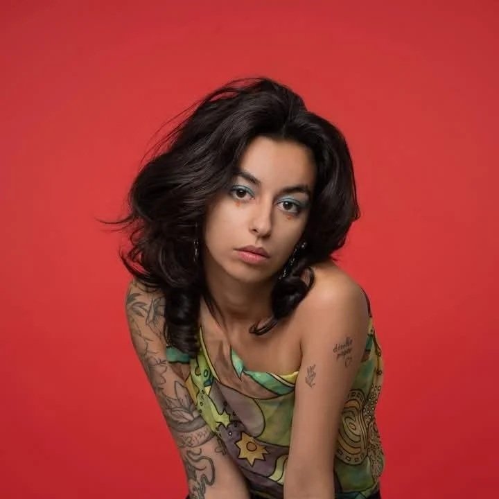 A young woman with dark, wavy hair and tattoos on her arms and shoulders, wearing a colorful top, posing against a red background.