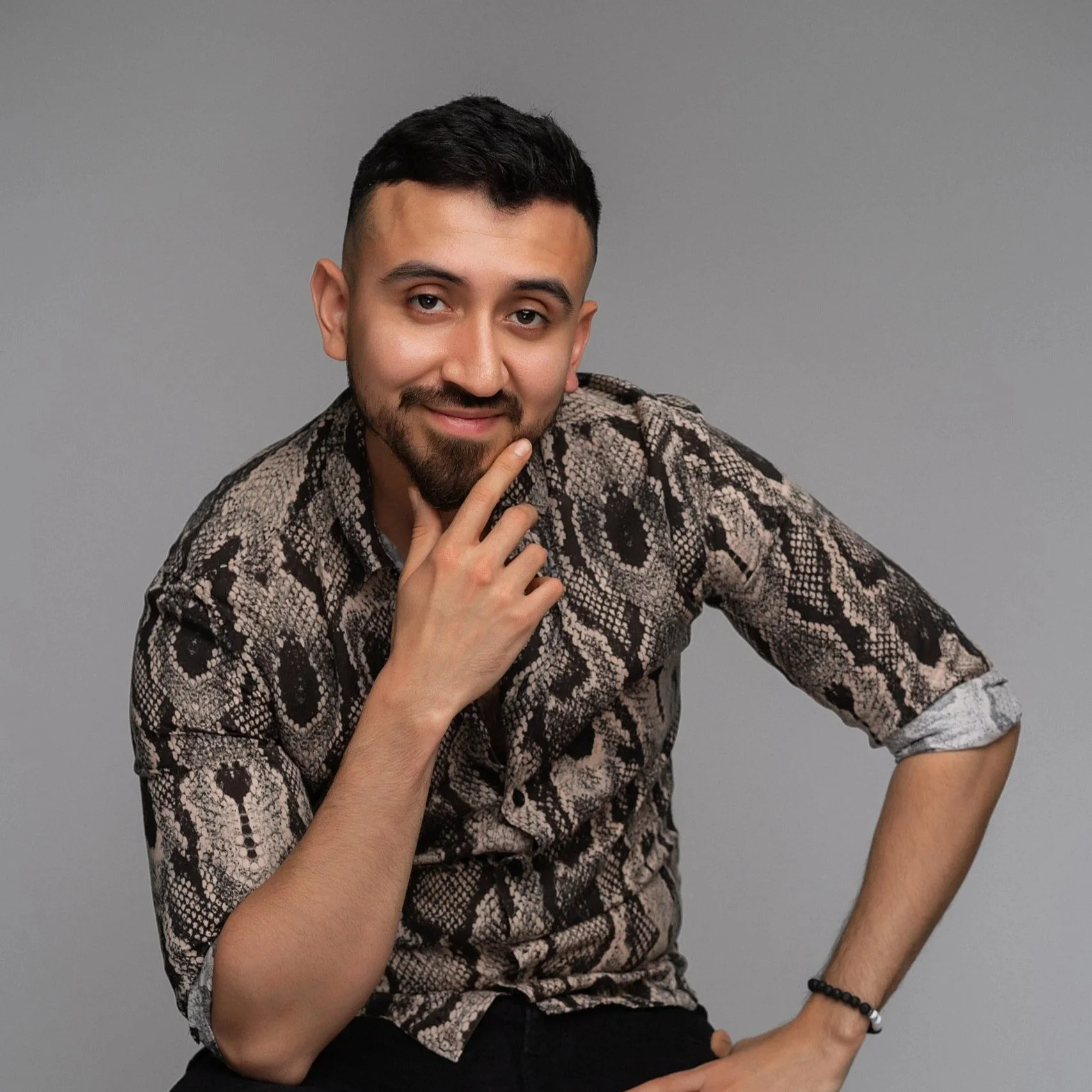A young man with dark hair and a beard, wearing a snake print shirt, poses with a thoughtful expression, resting his chin on his hand against a gray background.