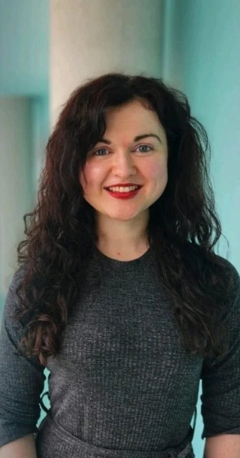 A woman with curly dark hair, wearing a dark gray top, smiling, standing indoors with a light green wall or curtain in the background.