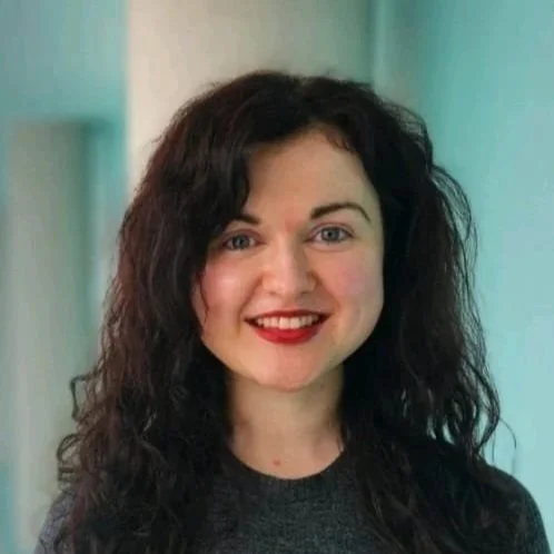 A woman with curly dark hair smiling at the camera, wearing a dark top, in an indoor setting with a light background.