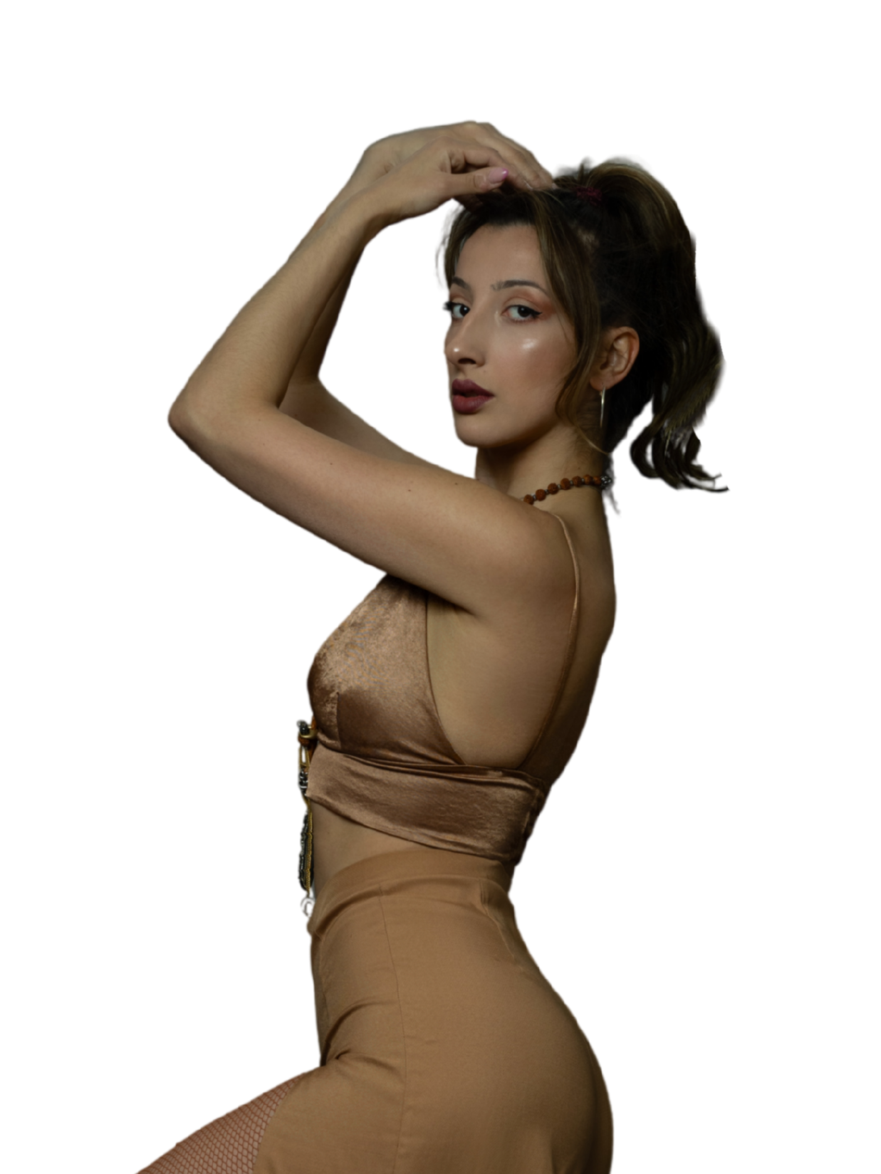 A woman with shoulder-length brown hair styled in waves, wearing a satin beige top and matching skirt, with one arm raised and hand resting on her head, posing against a black background.