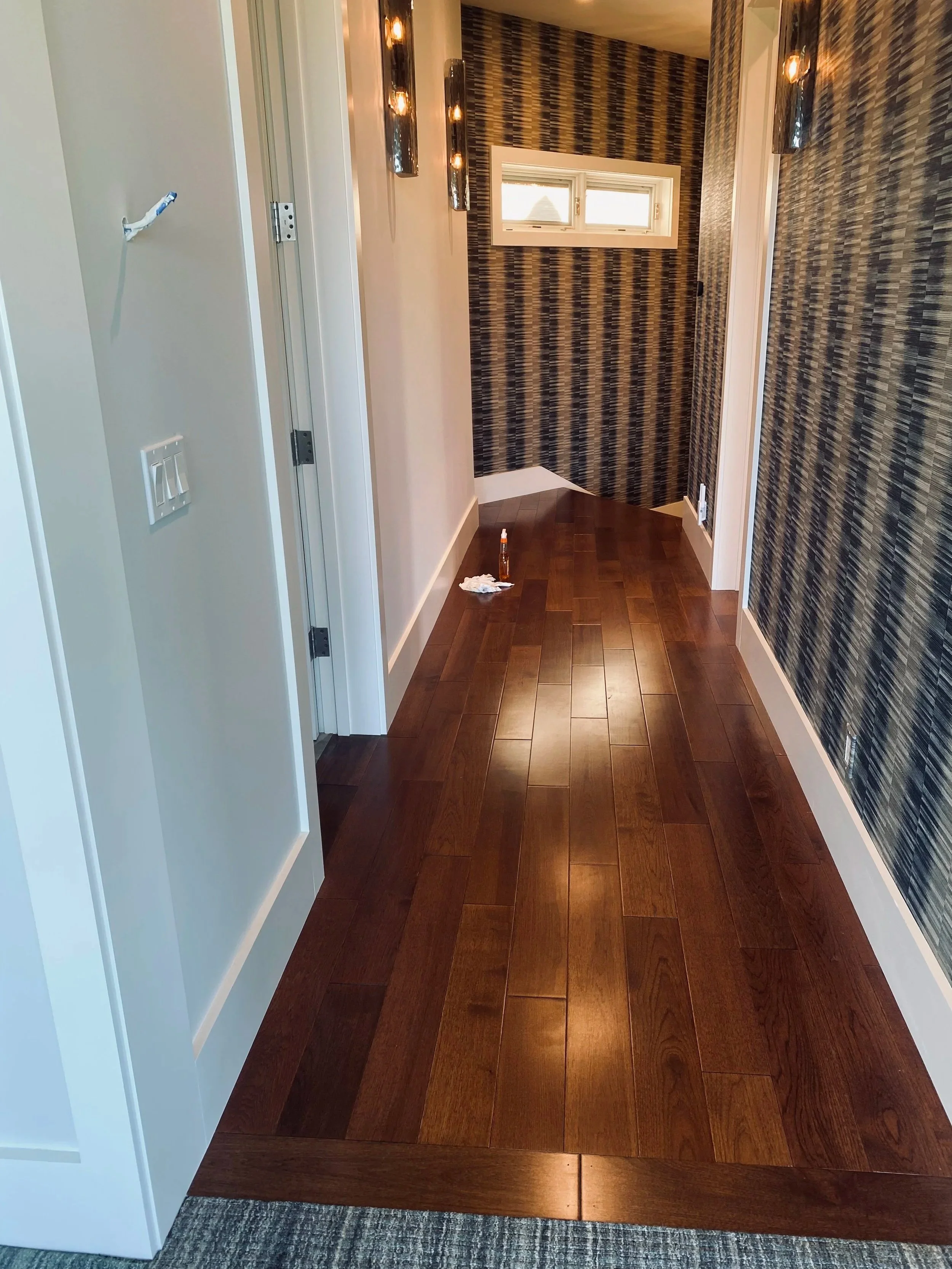 A hallway with dark wooden flooring, a white wall on the left, and a wall with a dark blue and gold patterned wallpaper on the right. In the distance, there is a small window and a corner with similar patterned wallpaper. A bottle and a cloth are on the floor near the left side of the hallway.
