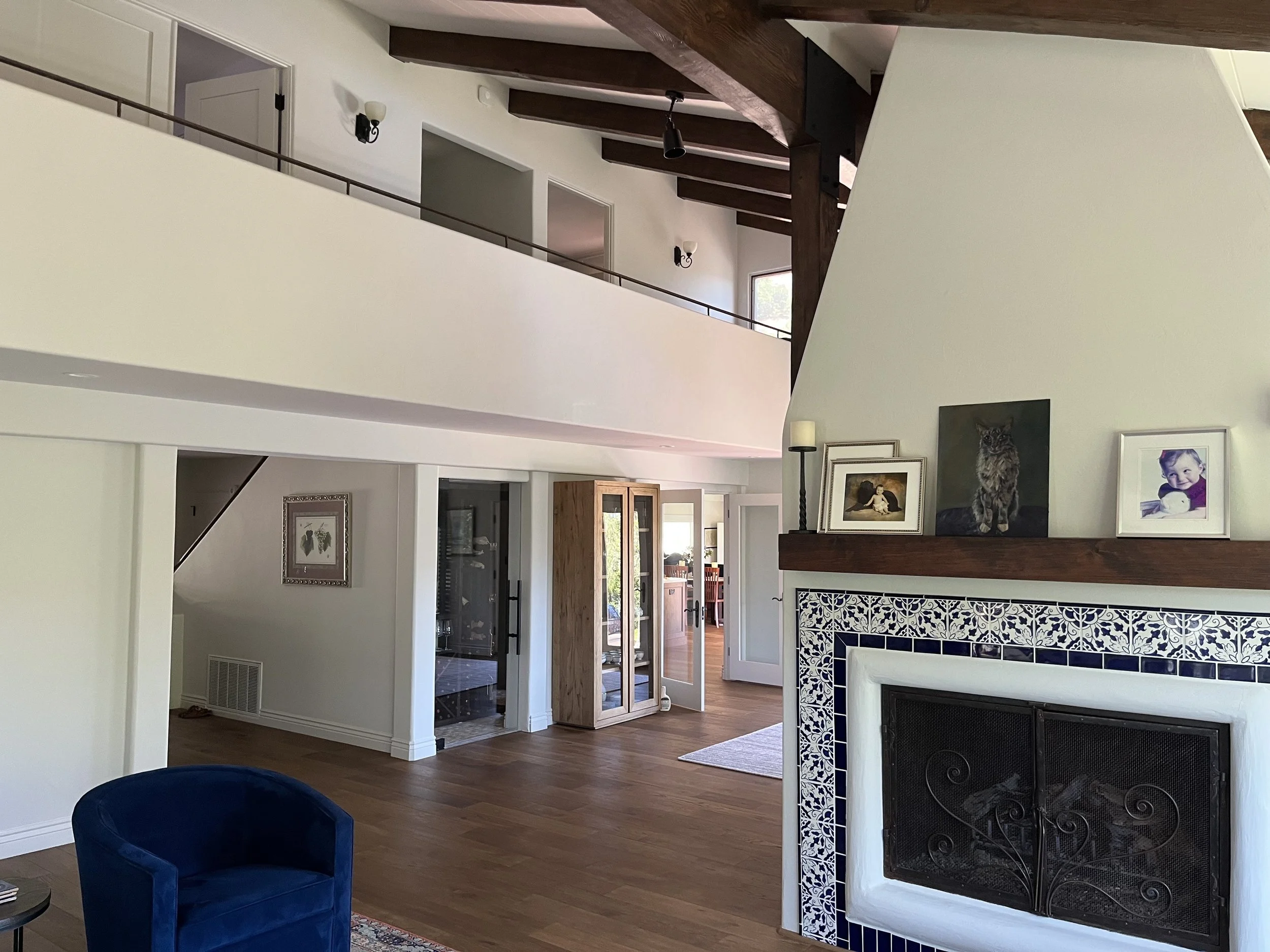 Interior of a modern home with a high ceiling, featuring a white wall fireplace with blue and white decorative tiles, and family photos on the mantle. There is a blue velvet chair, a wooden cabinet, and a cozy entryway with open doors leading to another room.