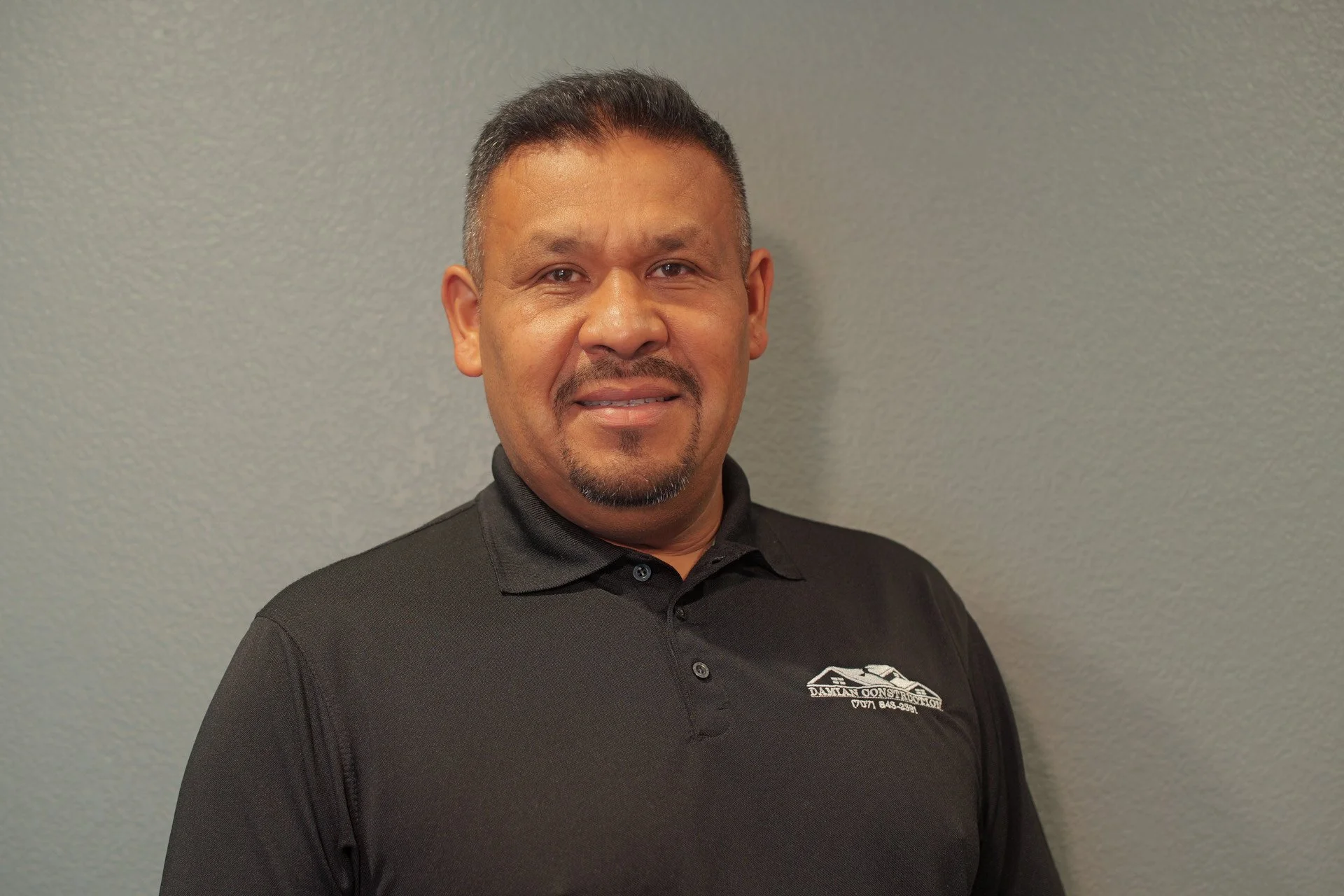 A man in a black collared shirt with a company logo standing against a light gray wall.