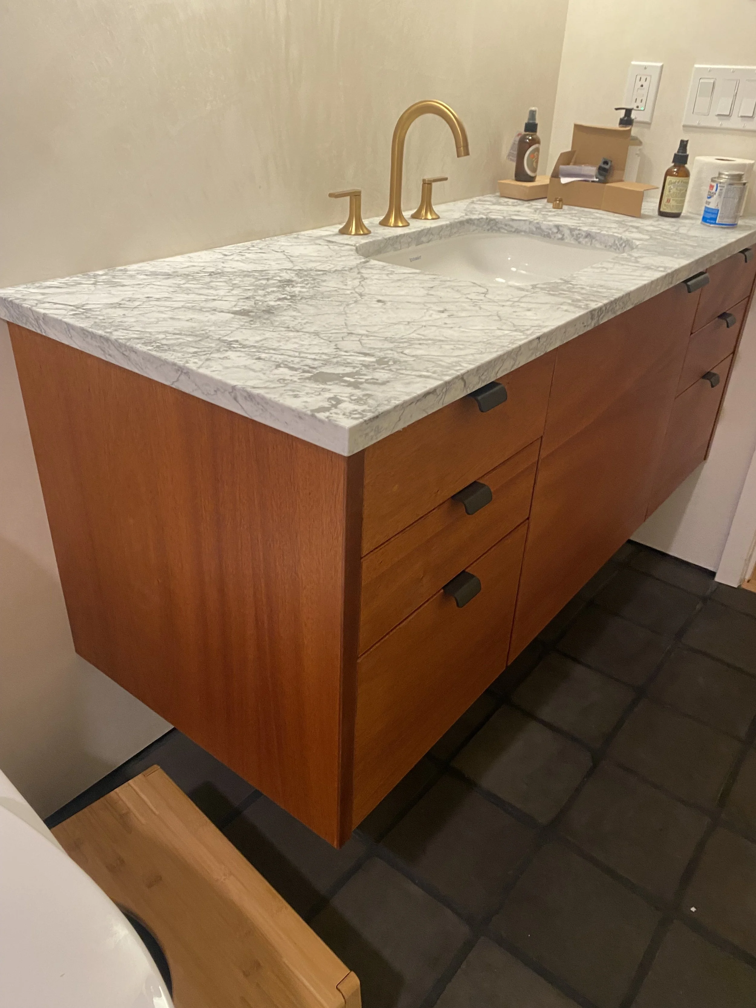 Bathroom vanity with a marble countertop, gold faucet and black drawer handles, against a beige wall with electrical outlets, and various toiletries on the countertop.
