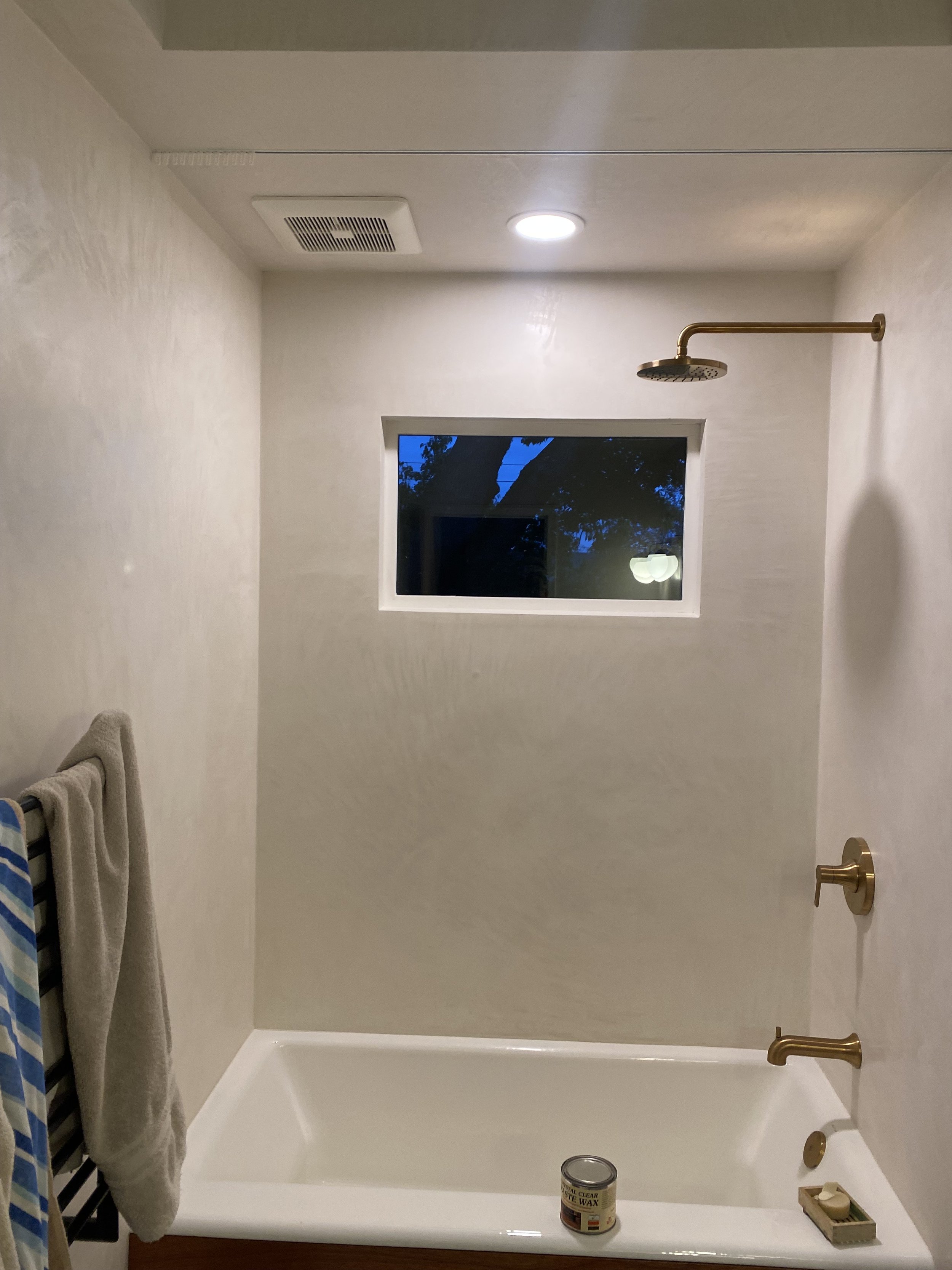 Bathroom shower with a window, beige walls, a brass showerhead, and a white bathtub. There is a towel hanging on a rack to the left and a small container of wax on the bathtub edge.