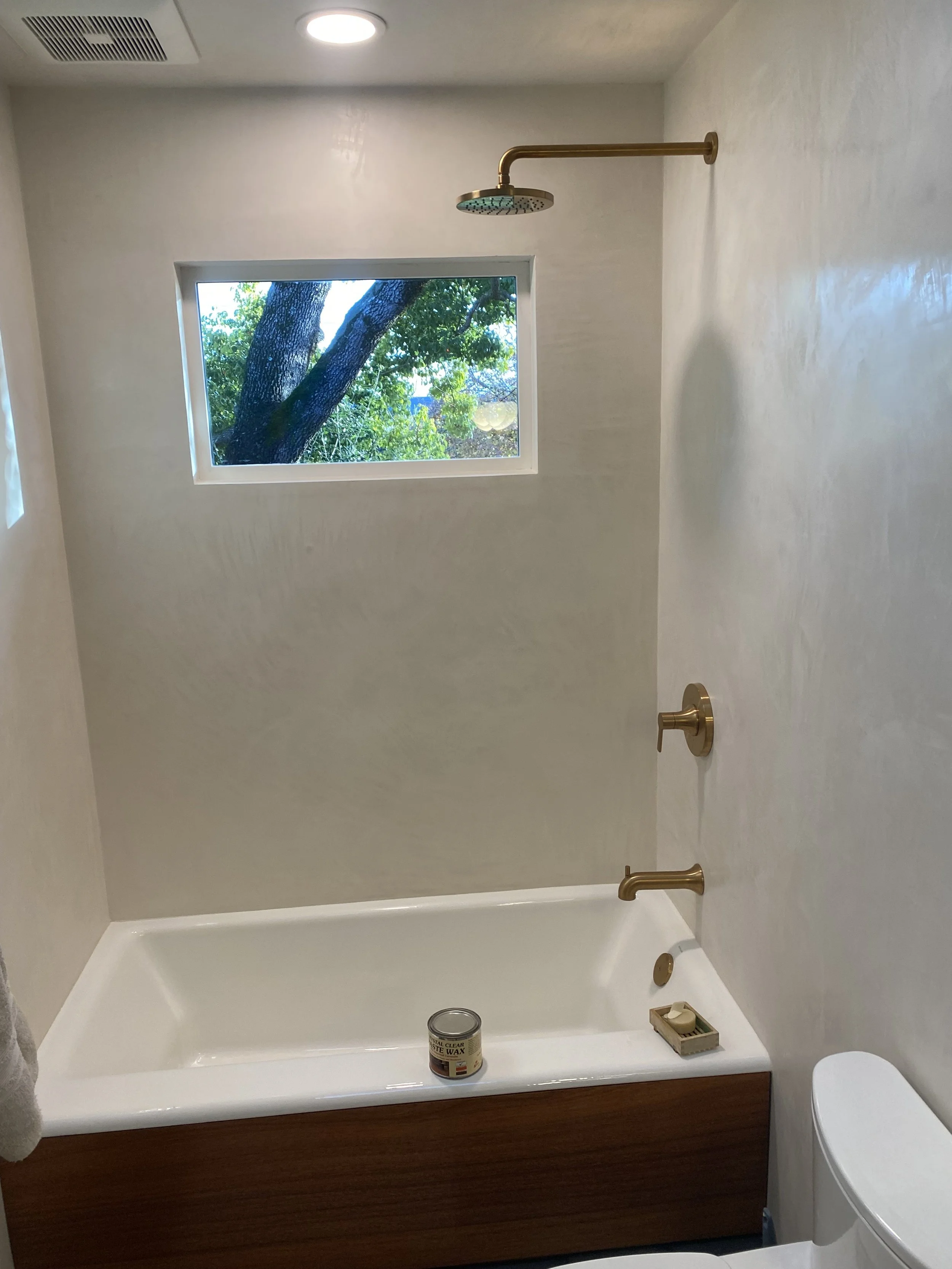 Bathroom with a bathtub, gold showerhead, and fixtures, small window showing green trees outside, and a can of wax on the tub ledge.