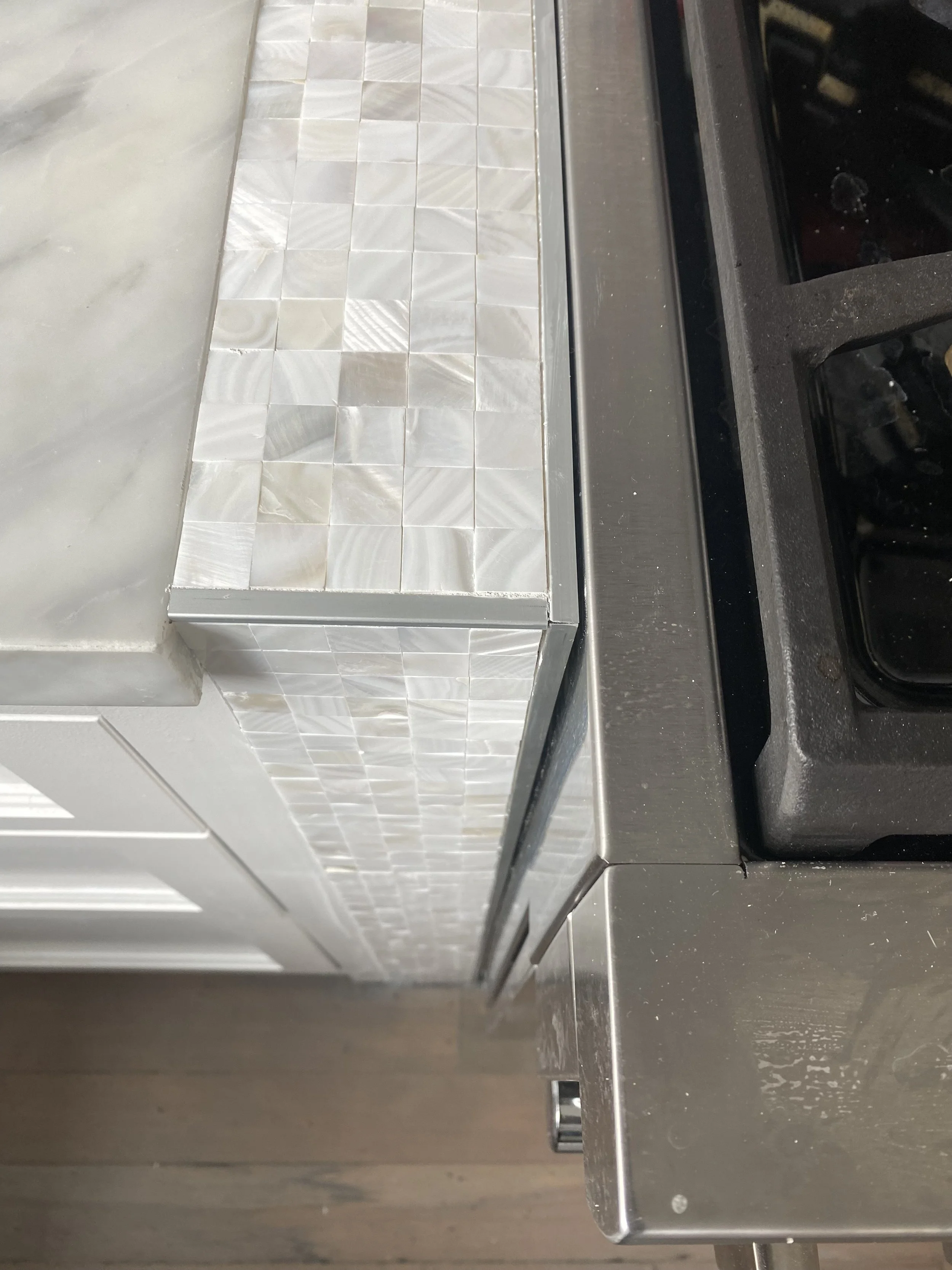 Close-up of a kitchen countertop with tiled marble and pearl-colored surface, next to a black stovetop on a grey metal counter.