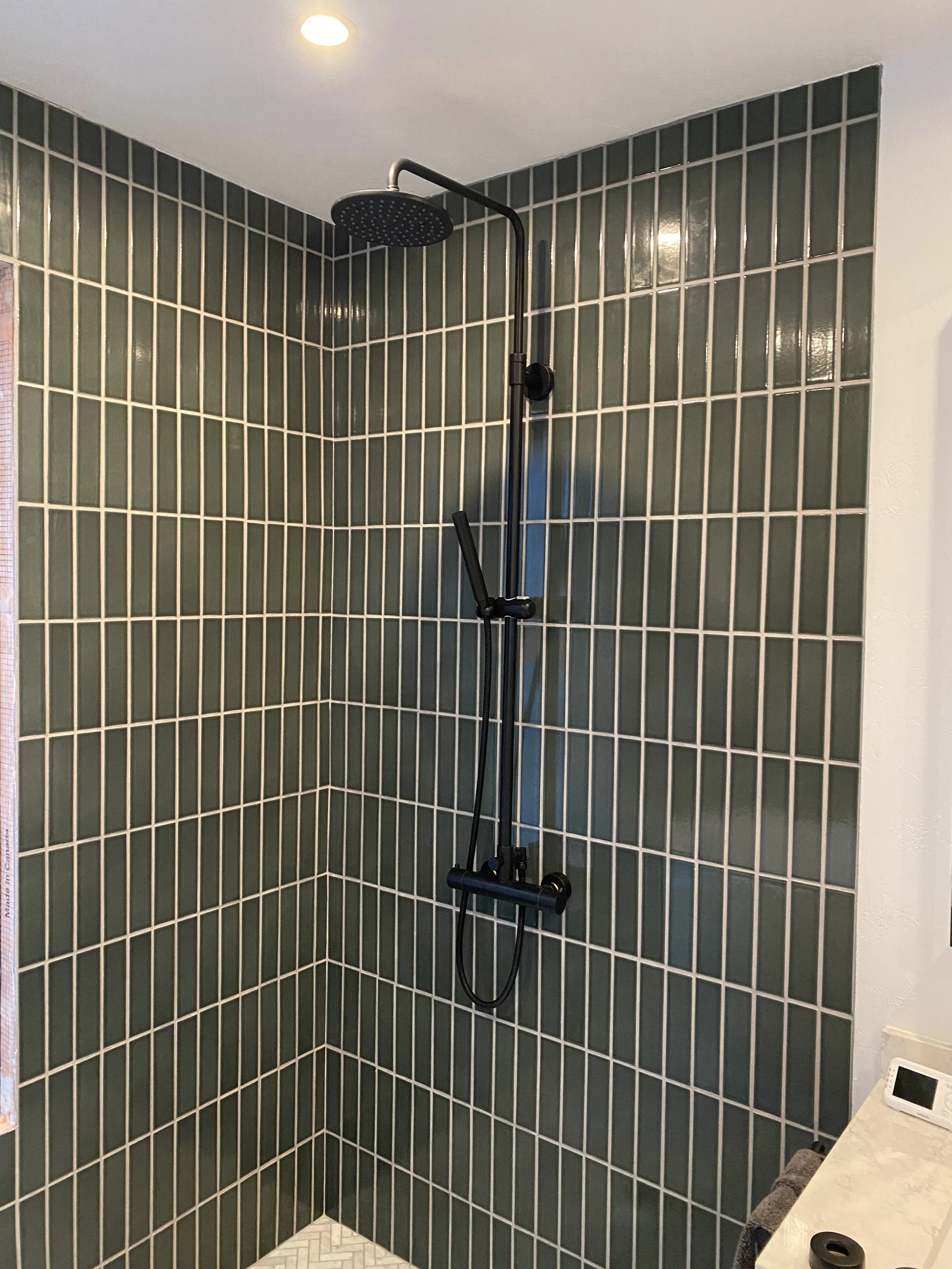Modern shower with black showerhead and handheld shower wand, green tiled wall, and white tiled floor.