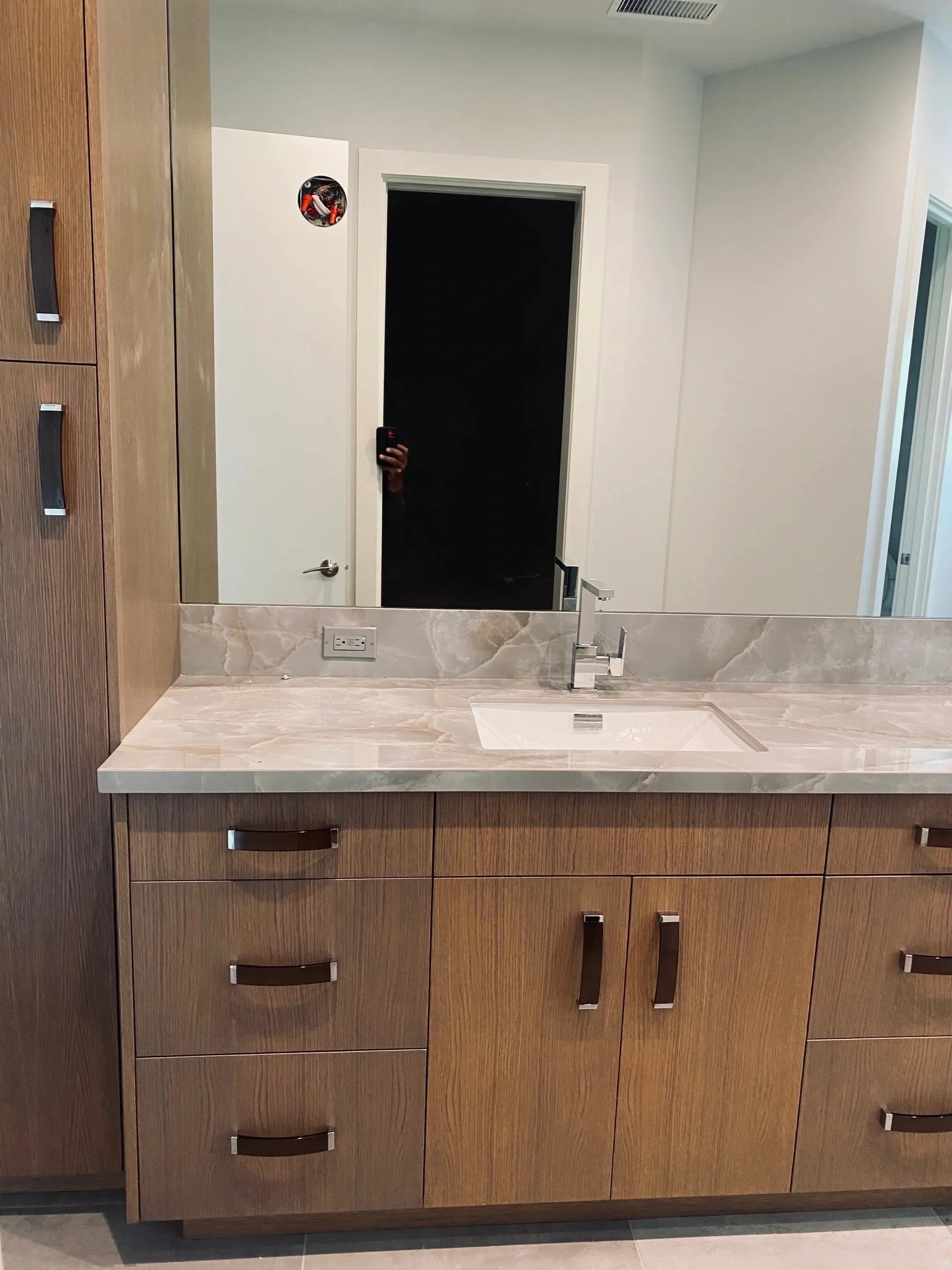 Bathroom vanity with a wooden cabinet, marble countertop, and a rectangular sink. Mirror above the sink and a wall outlet, with a partially visible door in the background.