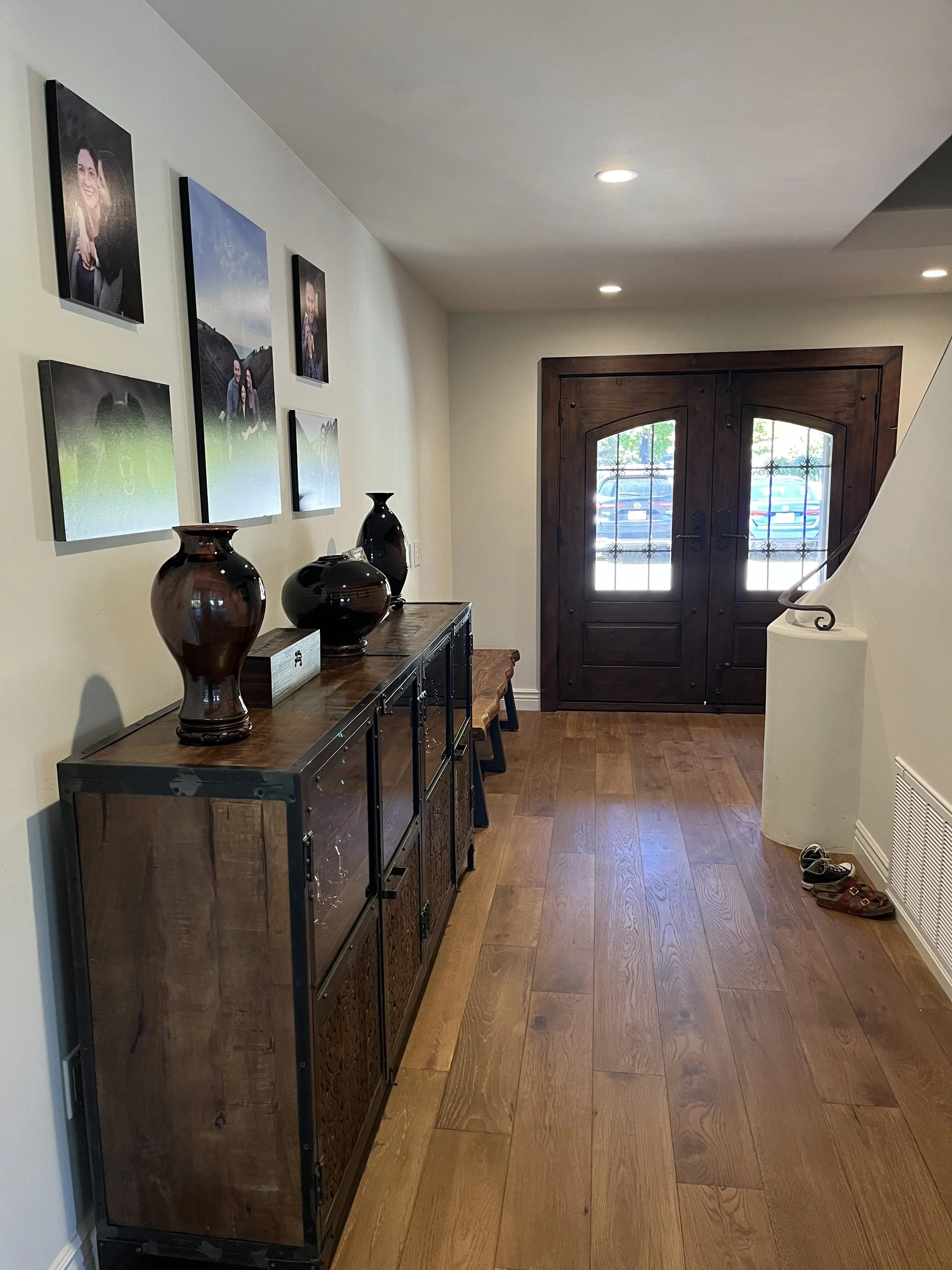 Entryway with wooden front door, hardwood floor, photo gallery on wall, dark wooden sideboard with vases, shoes on floor.