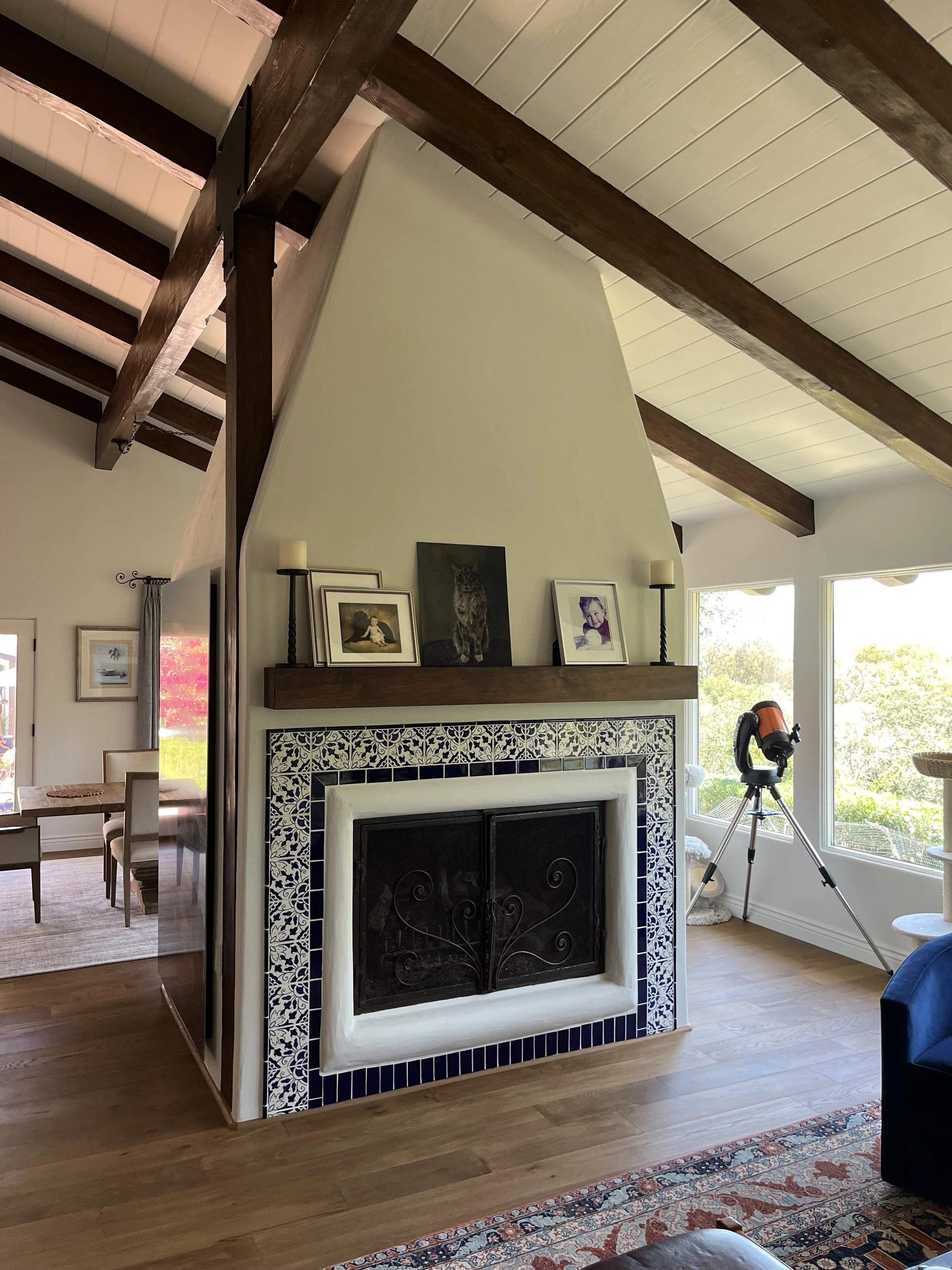 A small corner of a living room with a fireplace with decorative blue and white tiles, a wooden mantel with framed photos and paintings, and a telescope set up by the window looking outside at trees.
