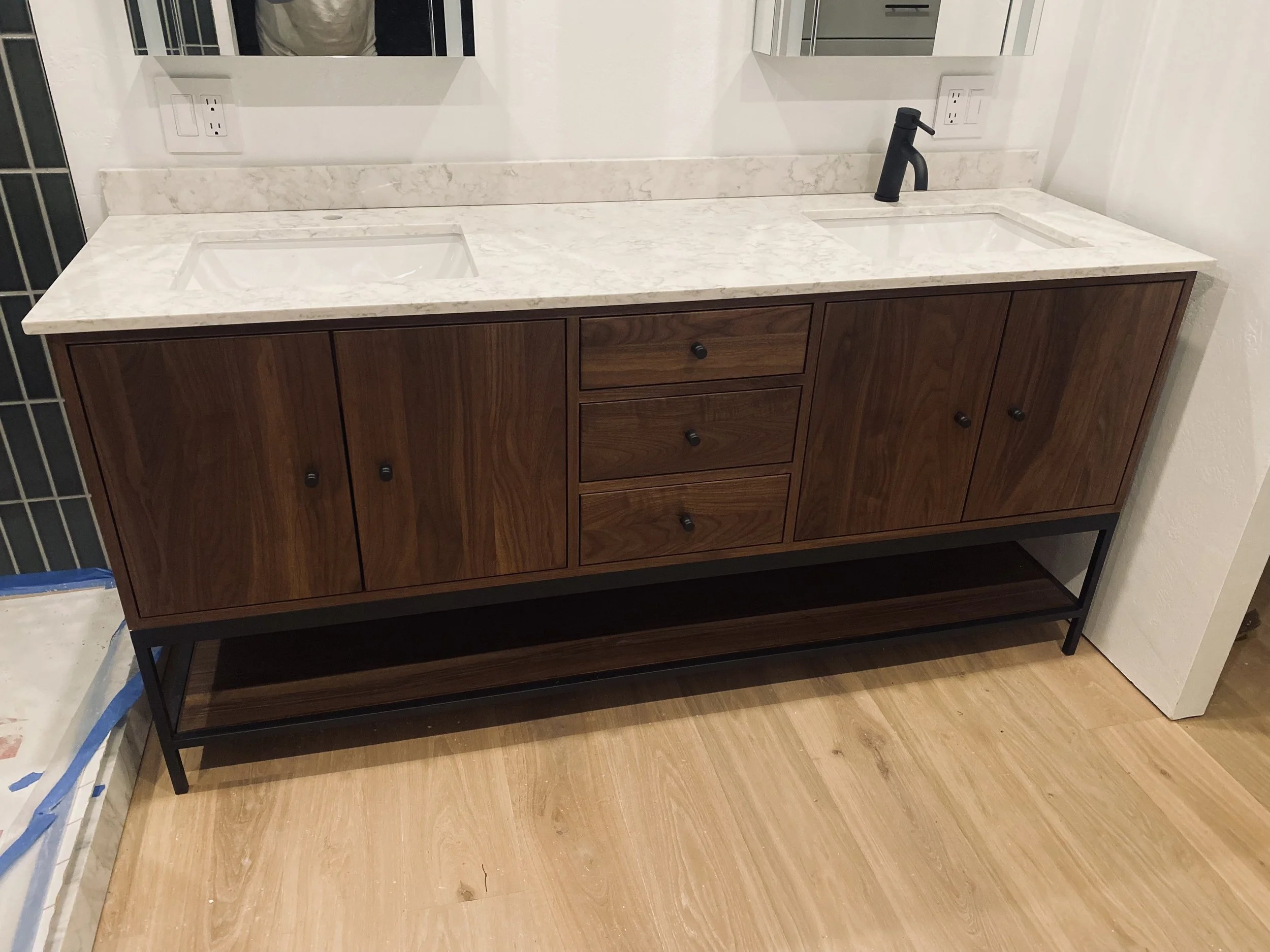 Modern bathroom vanity with a white marble countertop, two sinks, and black fixtures, featuring wooden cabinets and an open shelf below, against a white wall with mirrors.