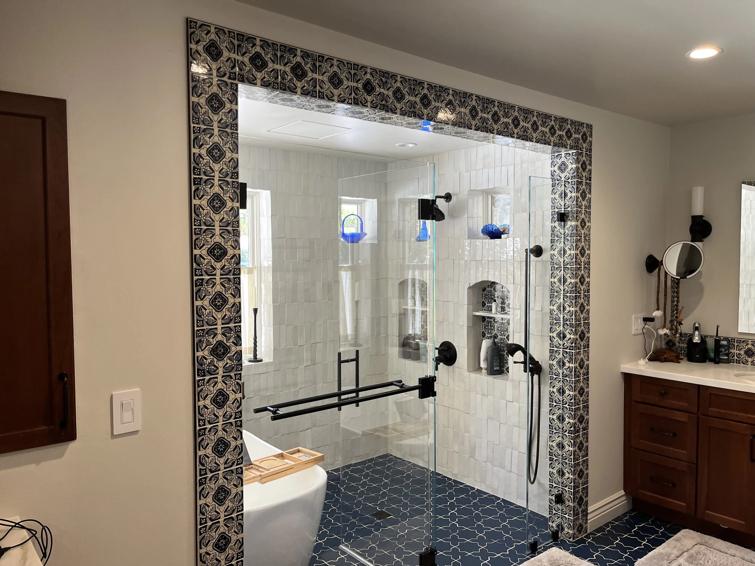 Bathroom with walk-in shower enclosed by glass, decorated with navy and white tiles, and a wooden vanity with a mirror and accessories nearby.