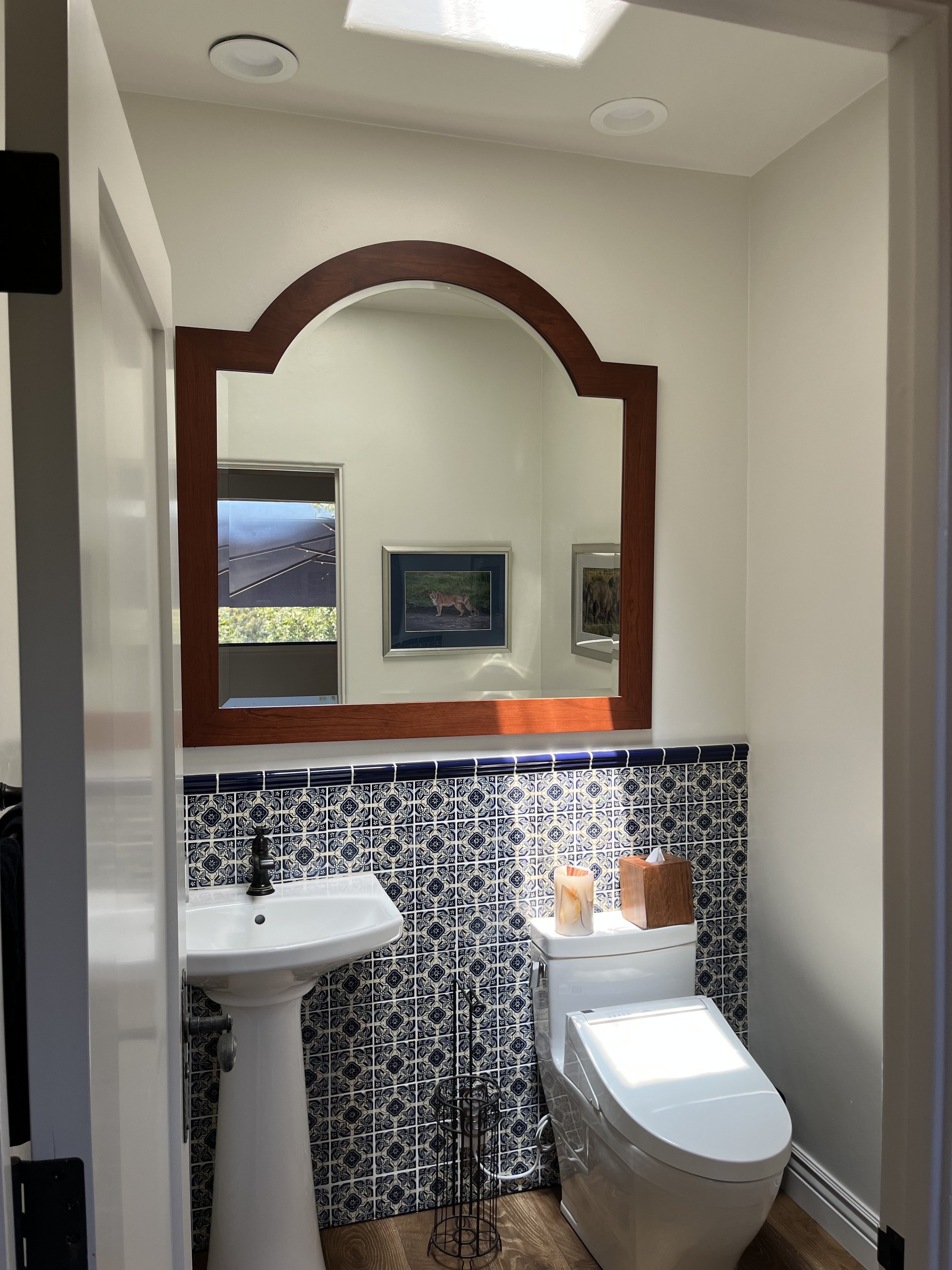A small bathroom with a pedestal sink, decorative tiled wall, a modern toilet, and a large wooden framed mirror. Small framed pictures are visible through the mirror's reflection. The room has a ceiling skylight and built-in ceiling lights.