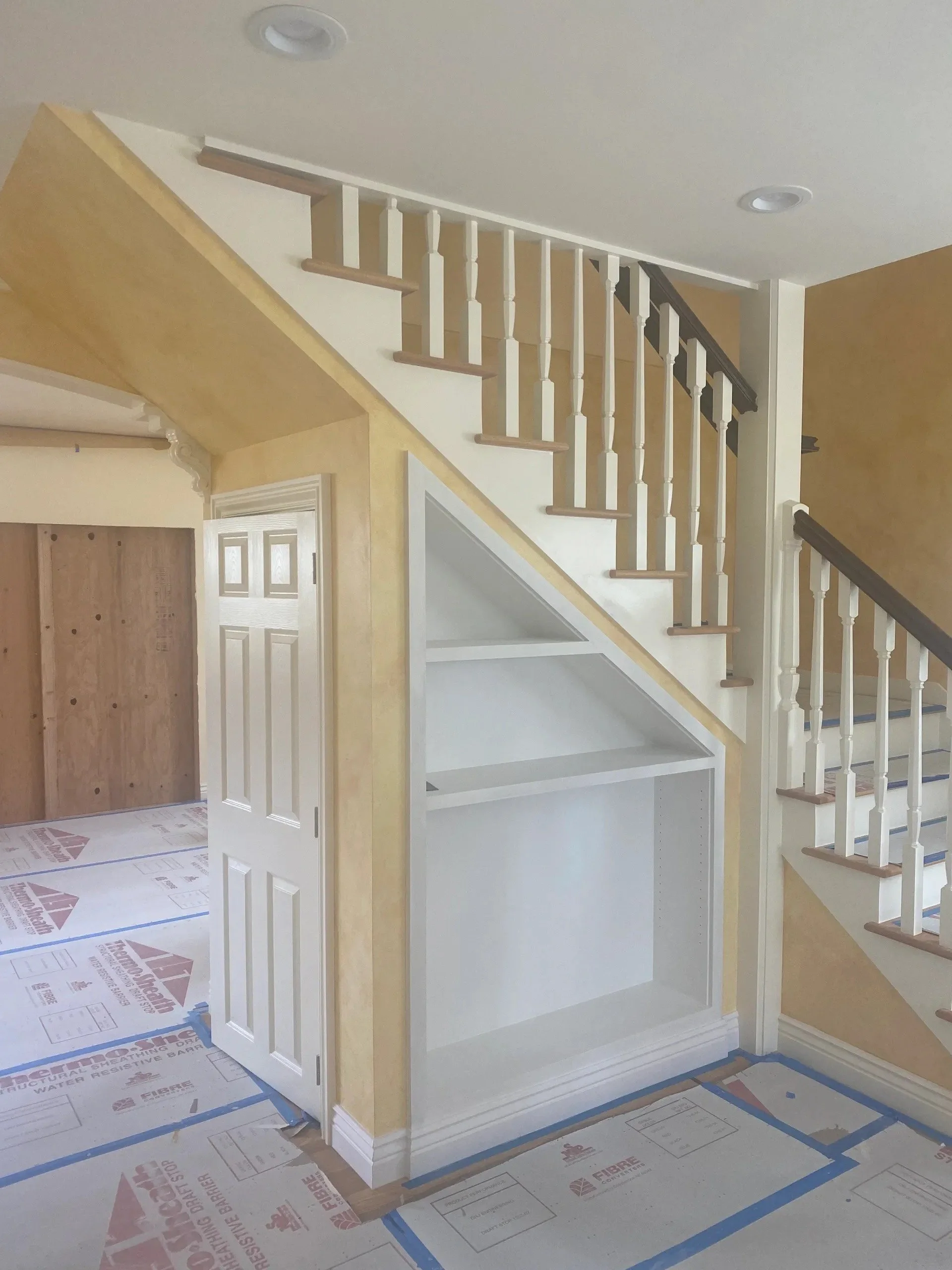 Interior view of a house under renovation showing a staircase, a small built-in shelving unit, and protective floor covering.