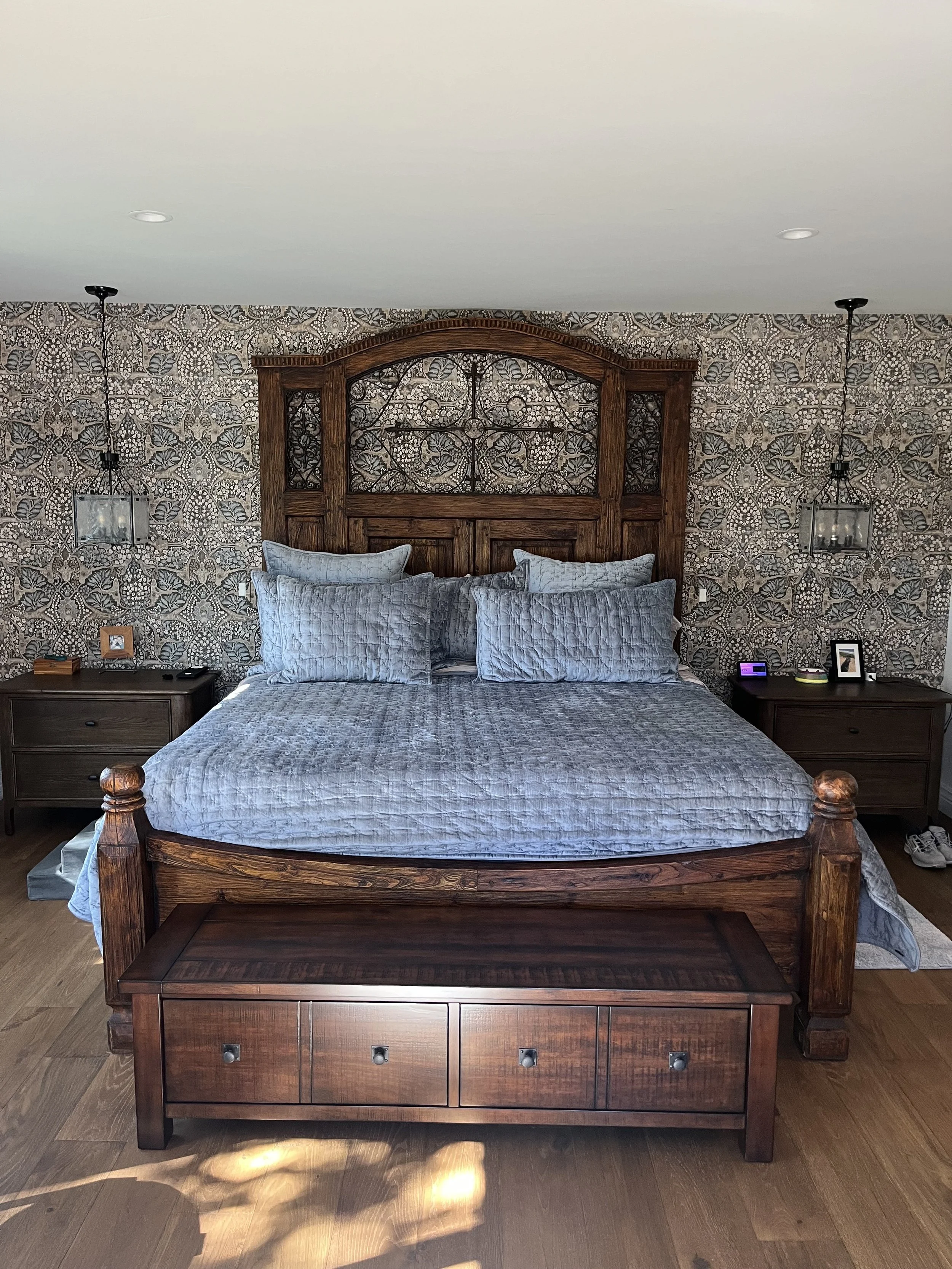 A bedroom with a large wooden bed featuring an ornate headboard, gray bedding, and matching pillows. There are two nightstands with lamps, clocks, and framed photos beside the bed. The wall behind has intricate patterned wallpaper, and there is a bench at the foot of the bed on a wooden floor.