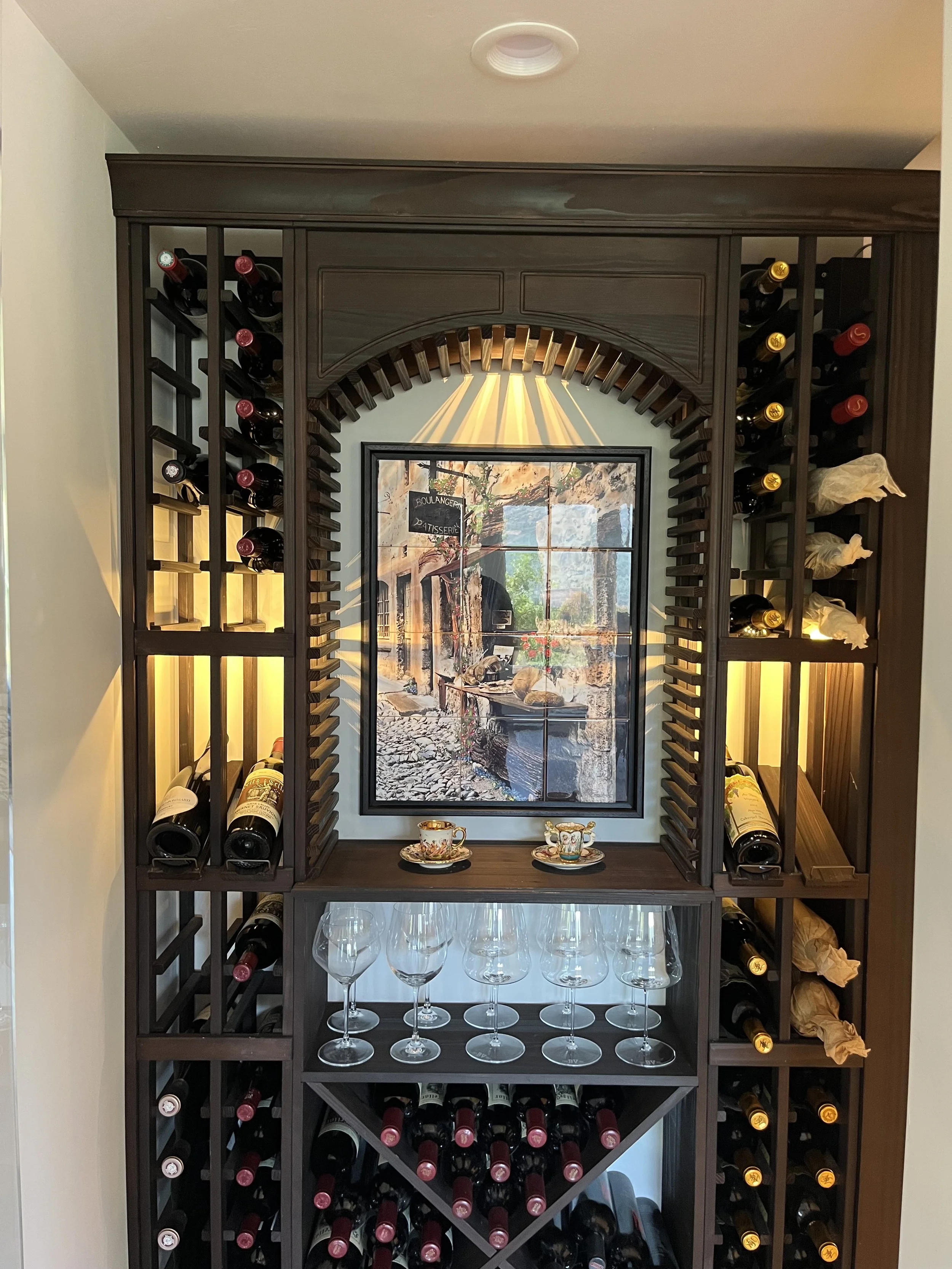 Dark wooden wine rack with bottles stored on the sides and bottom, decorative cups and a mirror with a landscape painting in the center.