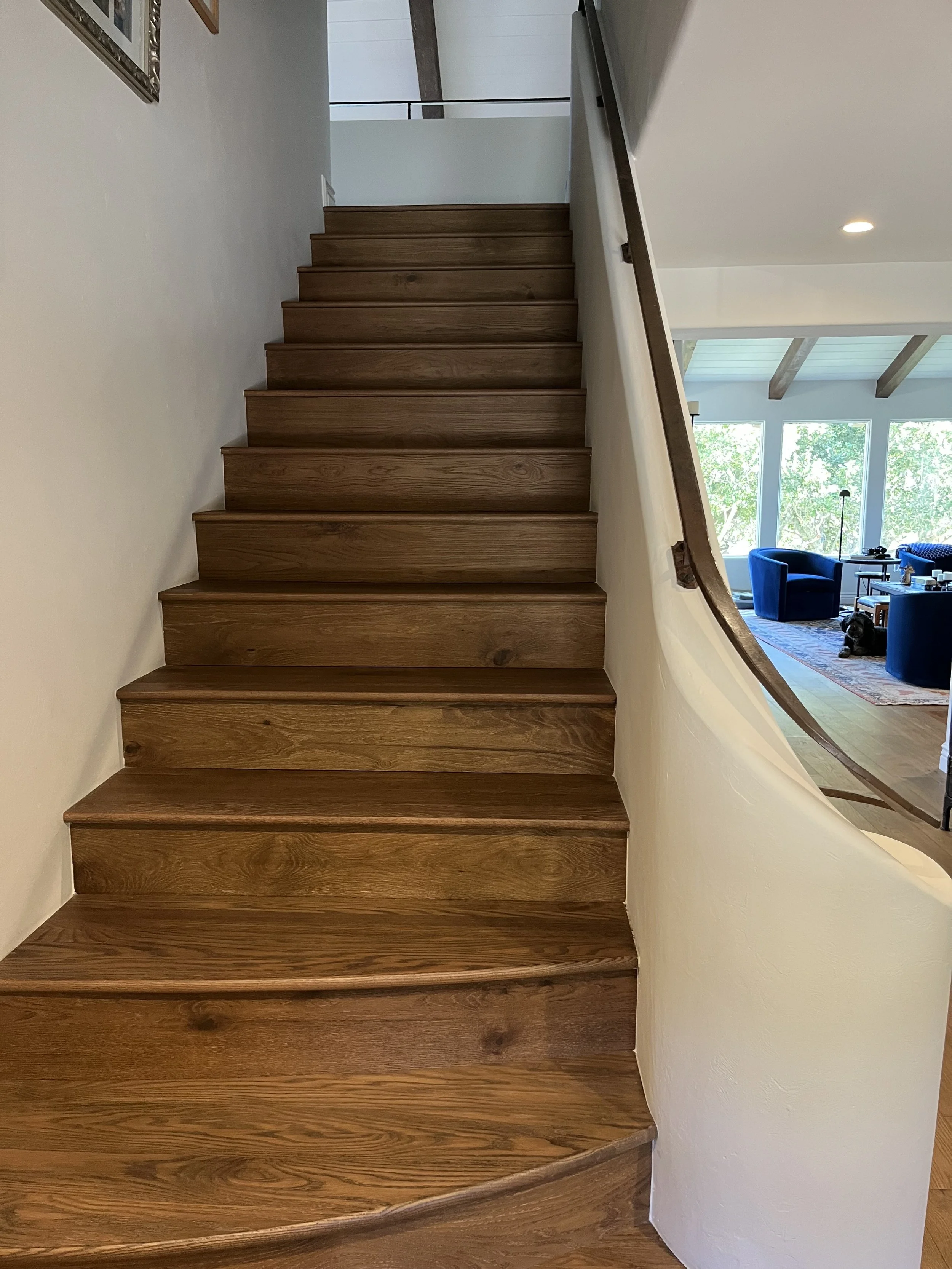 Wooden staircase inside a house, with a curved railing on the right side and a view into a living room with large windows and blue chairs.