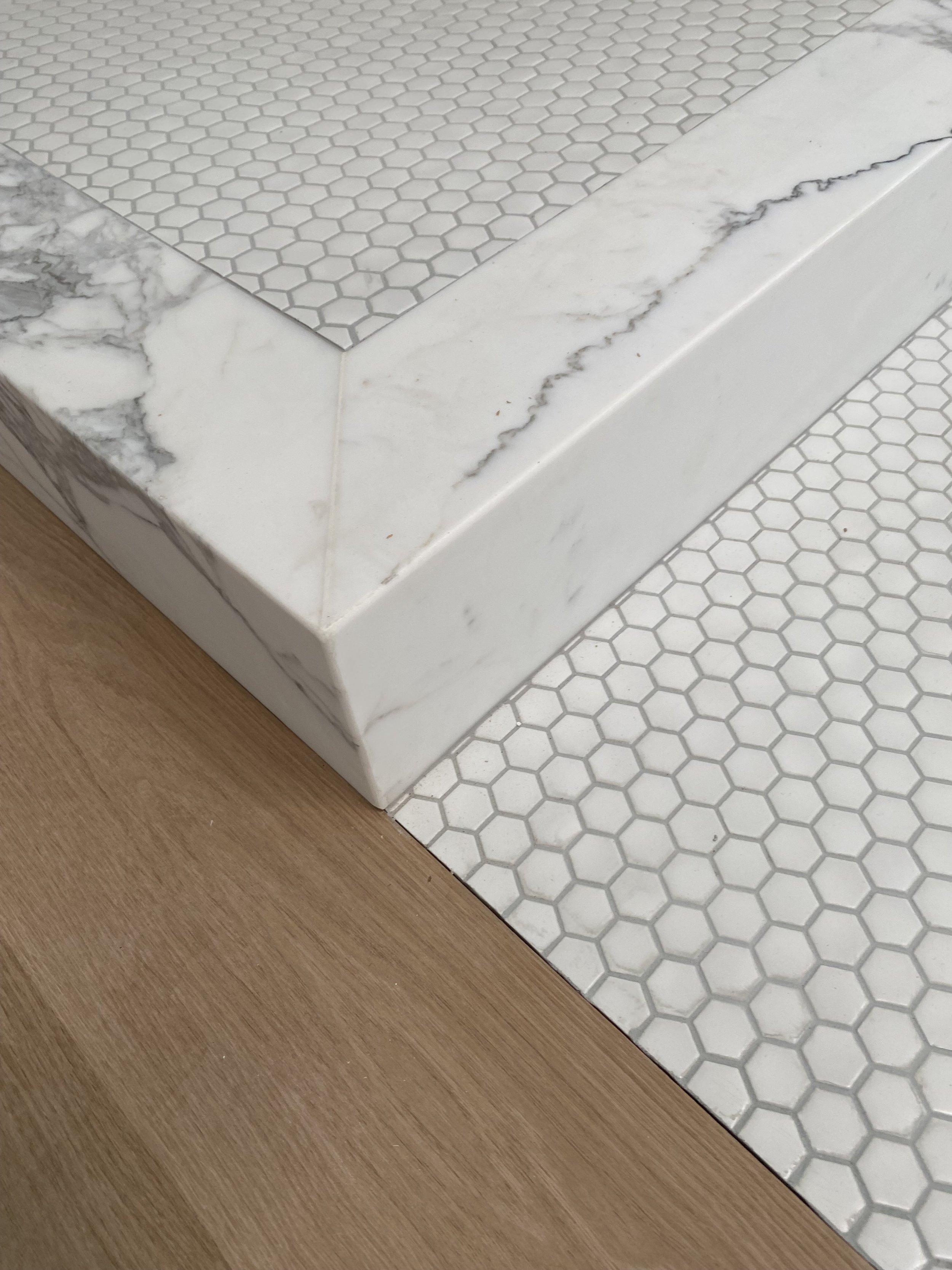 Close-up of a corner in a room showing white marble with gray veining on a raised platform, next to white hexagonal tile flooring, and a wooden floor.